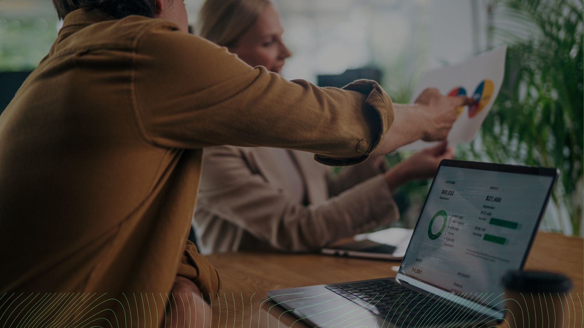 Two people working at a table with a laptop and printed report, analyzing financial data in an office with greenery outside.
