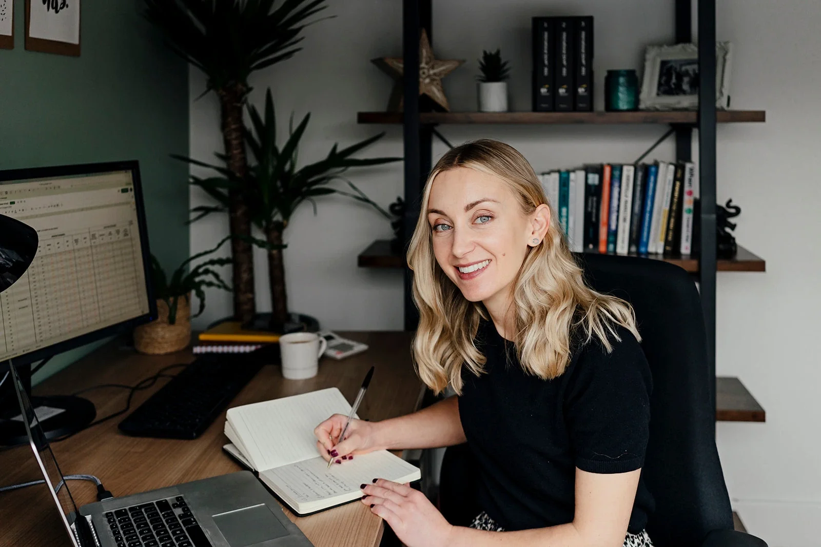 A woman working at her desk, writing in a notebook with a computer monitor, keyboard, and a mug nearby, in an office with bookshelves, plants, and decorative items in the background.