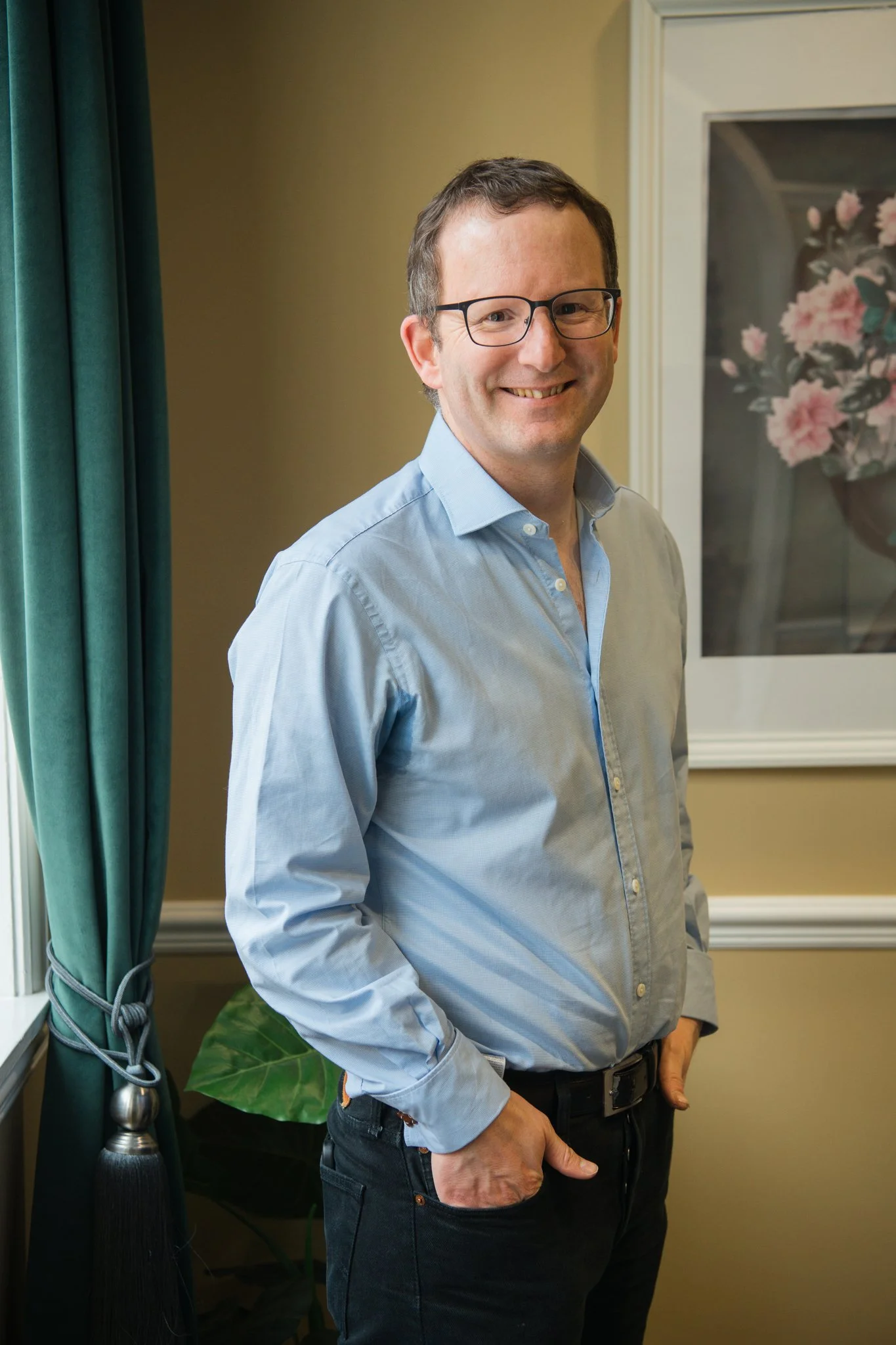A man with glasses and dark hair wearing a light blue button-up shirt and dark jeans standing indoors in front of a framed painting of pink flowers and green leaves, next to a window with teal curtains.