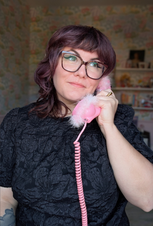 Une femme avec des lunettes, cheveux bouclés, tenant un téléphone rose avec du peluche, souriante, dans une pièce décorée de façon colorée.