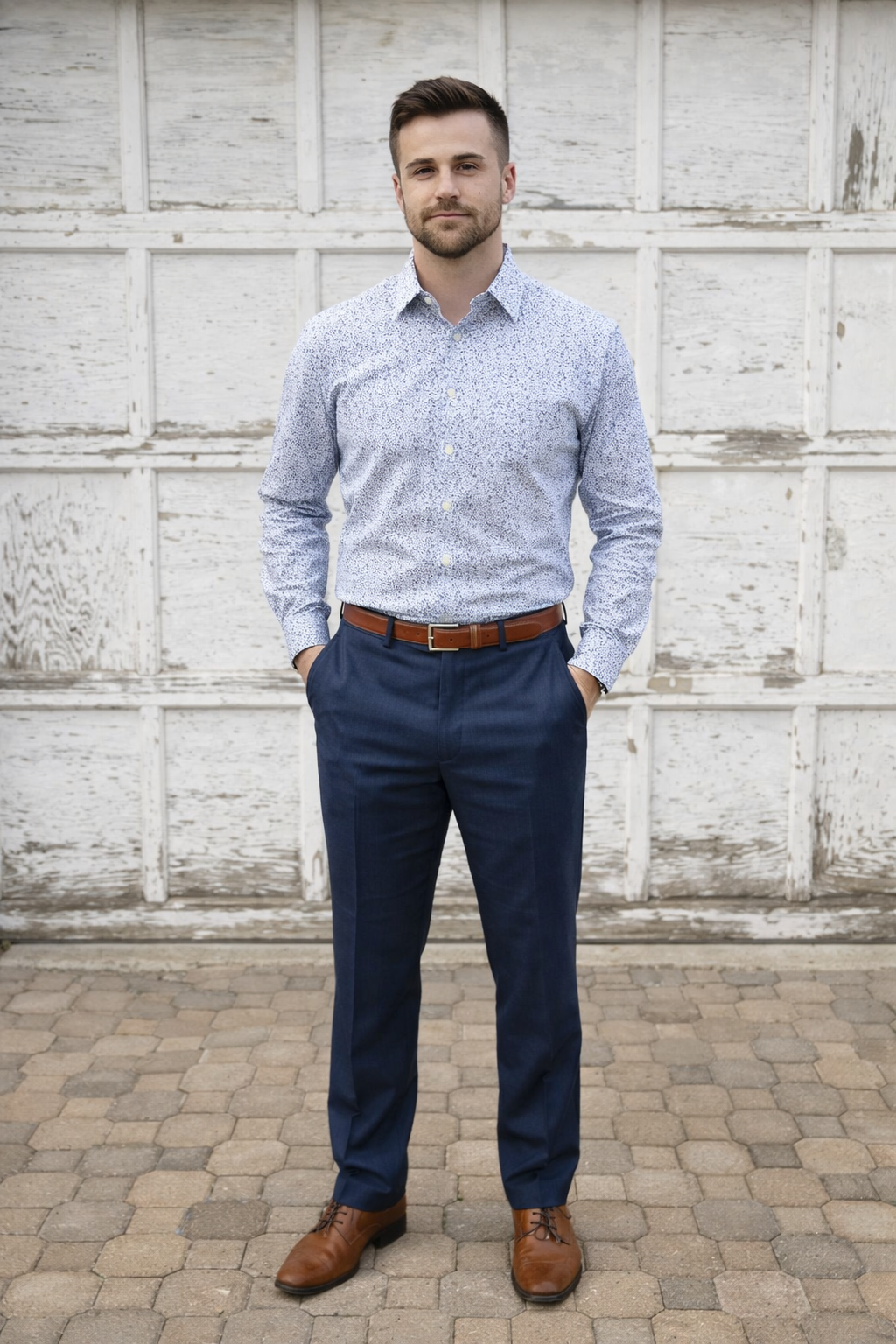 A man standing outdoors against a weathered white wooden wall, wearing a light blue patterned dress shirt, dark blue pants, brown belt, and brown dress shoes, with his hands in his pockets.
