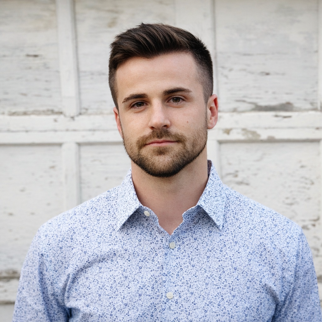 A young man with short dark hair and a beard, wearing a light blue patterned button-up shirt, posing outdoors in front of a white wooden wall.