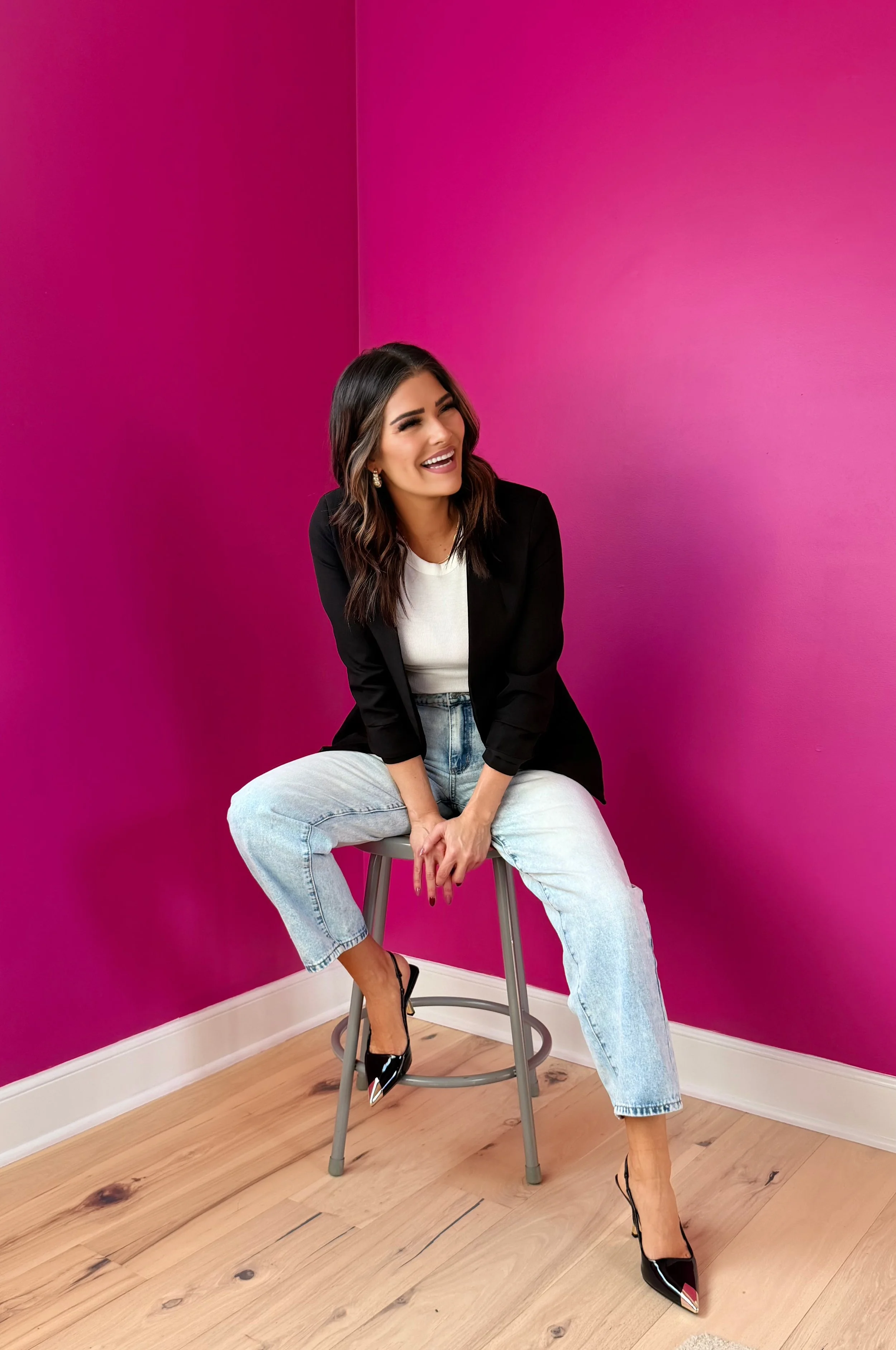 A woman with dark brown hair styled in loose waves, wearing a black blazer, white top, light blue jeans, and black high heels, sitting on a tall wooden stool with crossed legs, smiling and looking to her right, against a vibrant pink wall in a room with light wooden flooring.