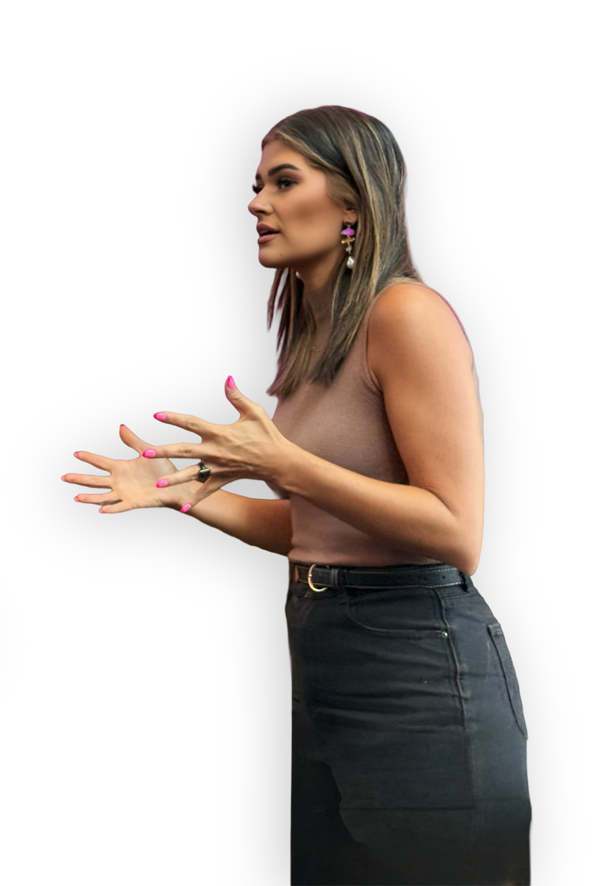 A woman with long, light brown hair with blonde highlights, wearing a beige sleeveless top and high-waisted black jeans, speaking and gesturing with her hands. She has pink earrings and pink nail polish.