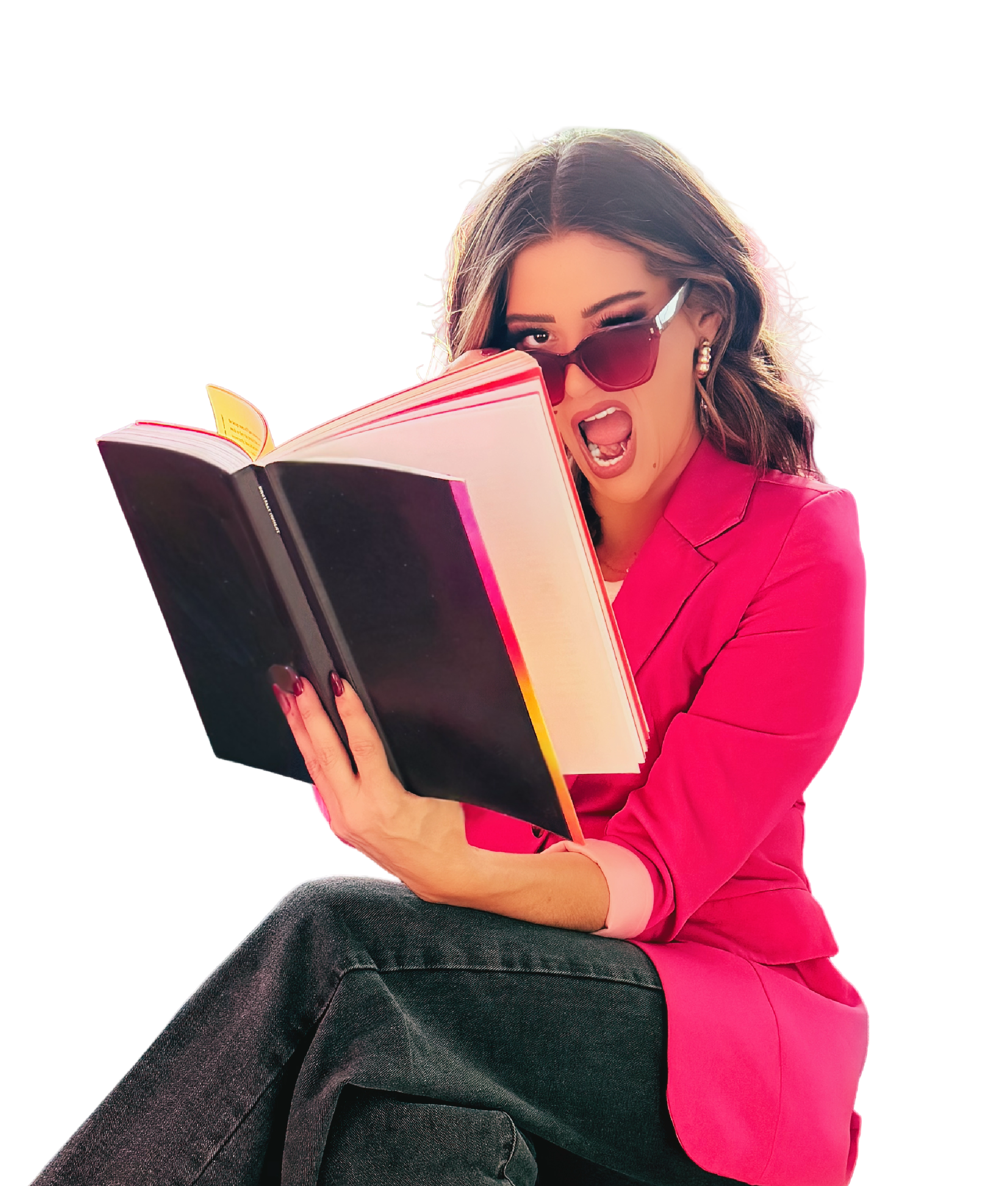 A woman with light skin, long wavy brown hair, wearing red sunglasses and a bright pink blazer, appears to be surprised or excited while reading a large book or magazine with colorful pages. She is sitting with her legs crossed, dressed in dark jeans, against a plain black background.