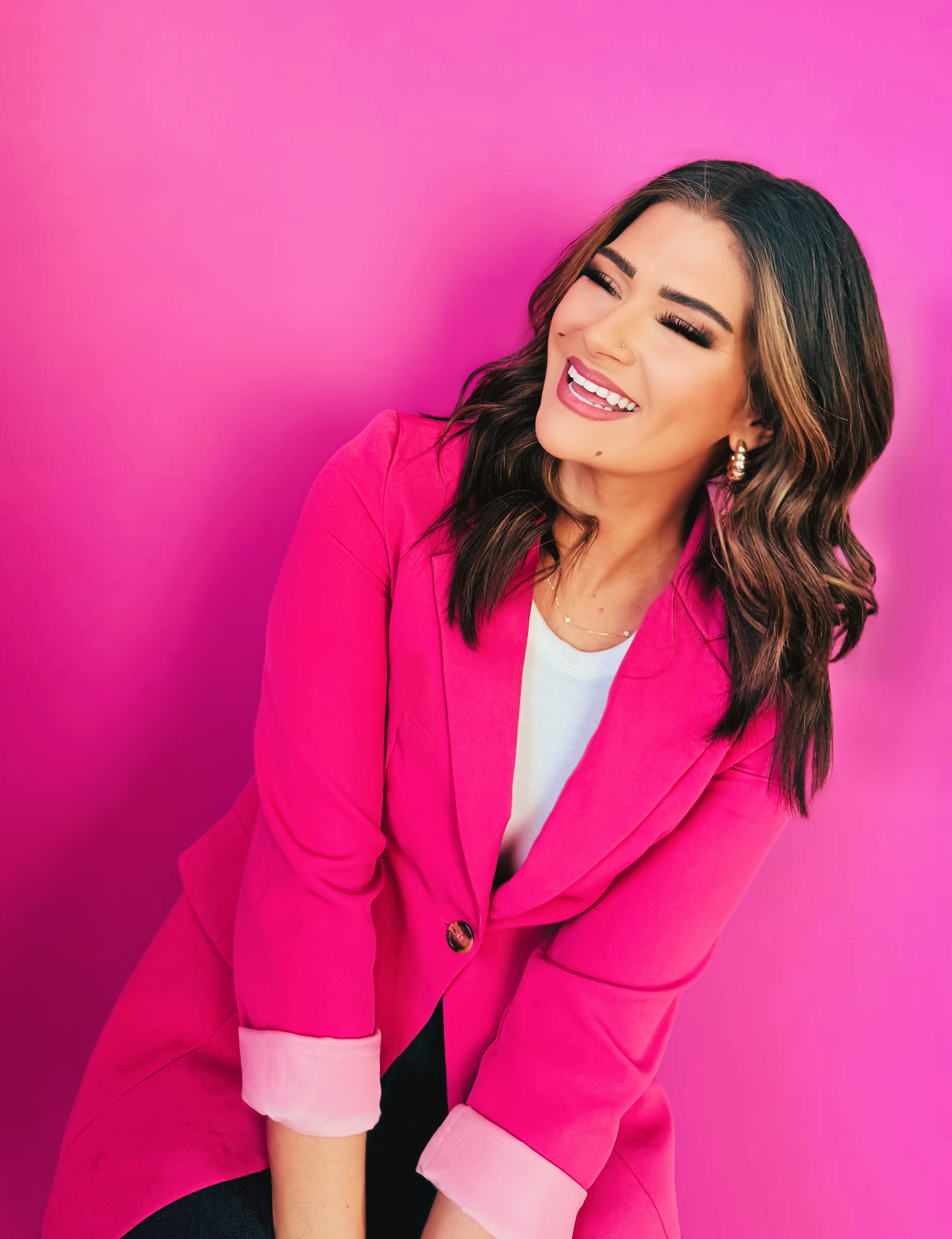 A woman wearing a bright pink blazer with rolled-up sleeves and a white shirt underneath, smiling and looking to the side, standing against a pink background.