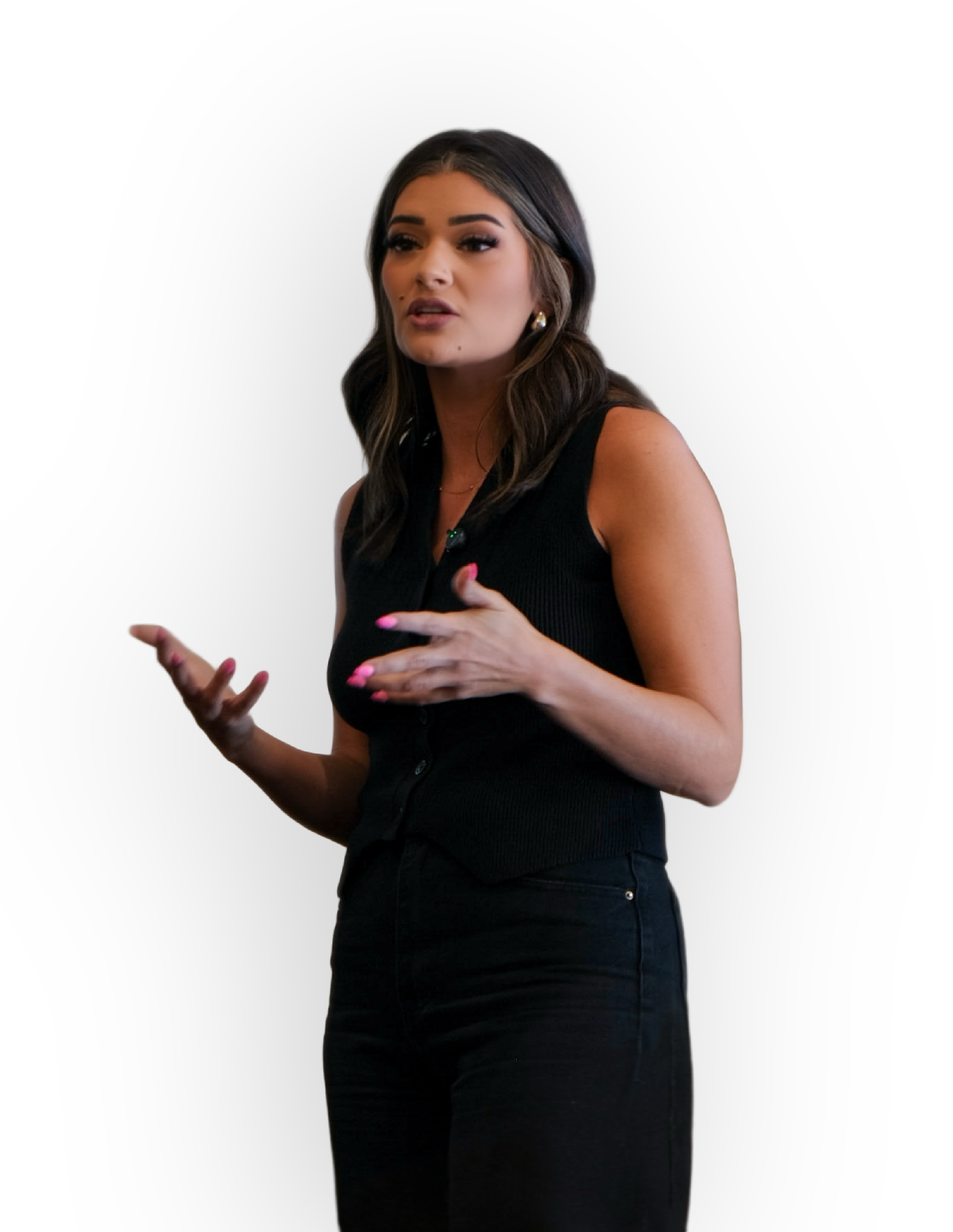 A woman with dark hair and makeup, dressed in a sleeveless black top and pants, speaking or gesturing with her hands.