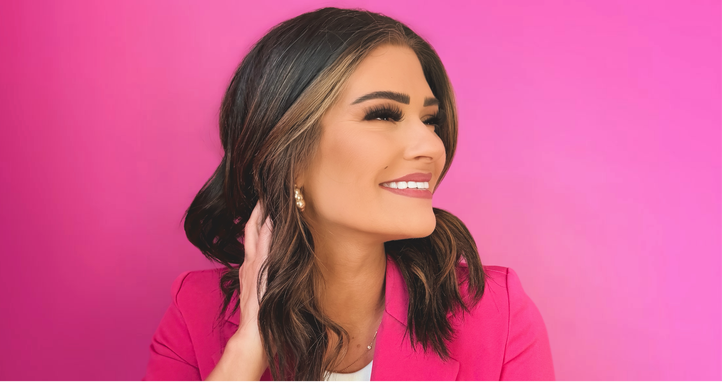 A woman with dark brown hair and blonde highlights smiling and looking to her right, wearing a pink blazer with a pink background.