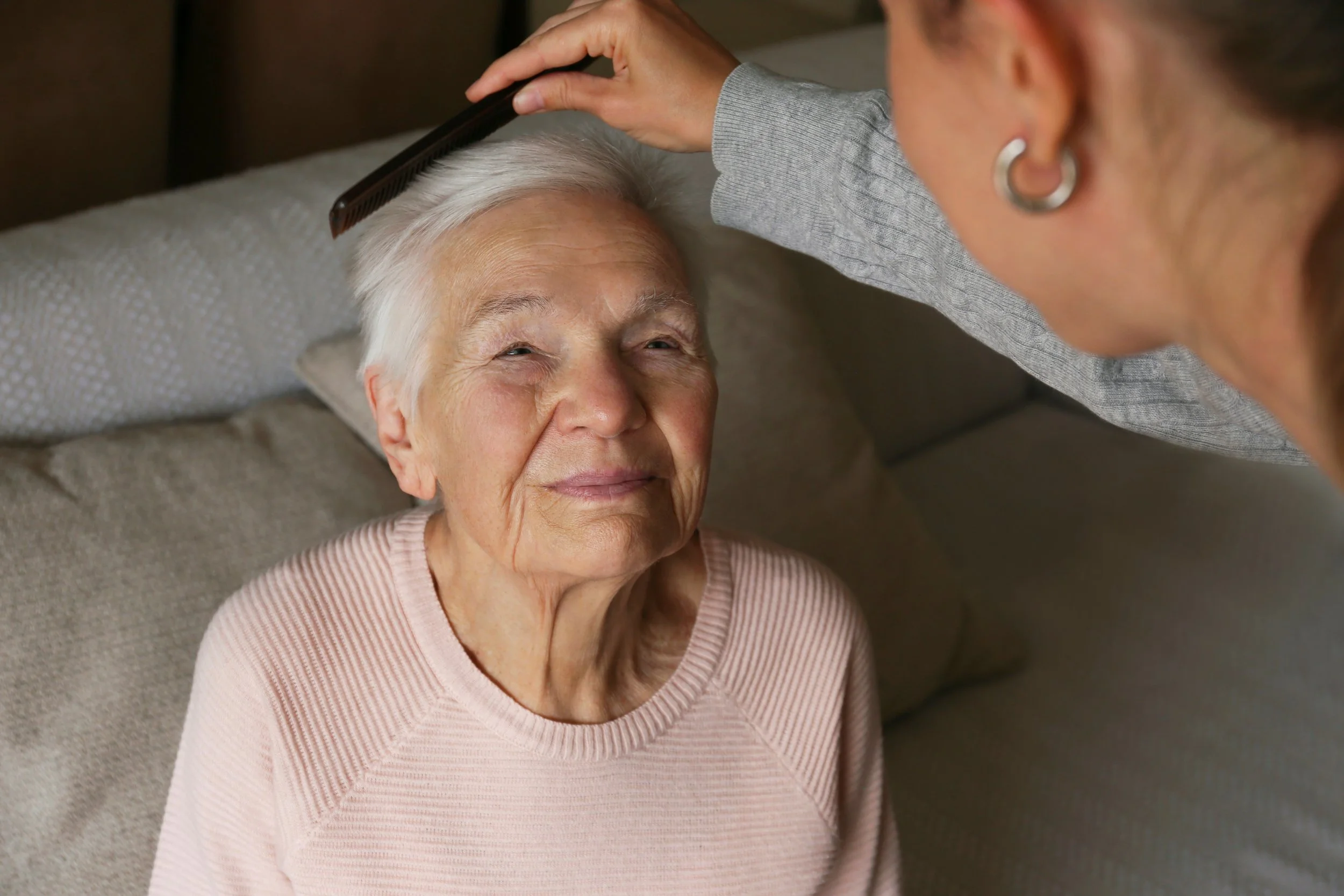 A woman brushes the hair of an elderly woman who is sitting on a couch, smiling gently.