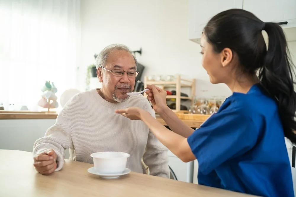 A woman in a blue shirt feeding a spoonful of soup or pudding to an elderly man seated at a table in a bright, modern kitchen.