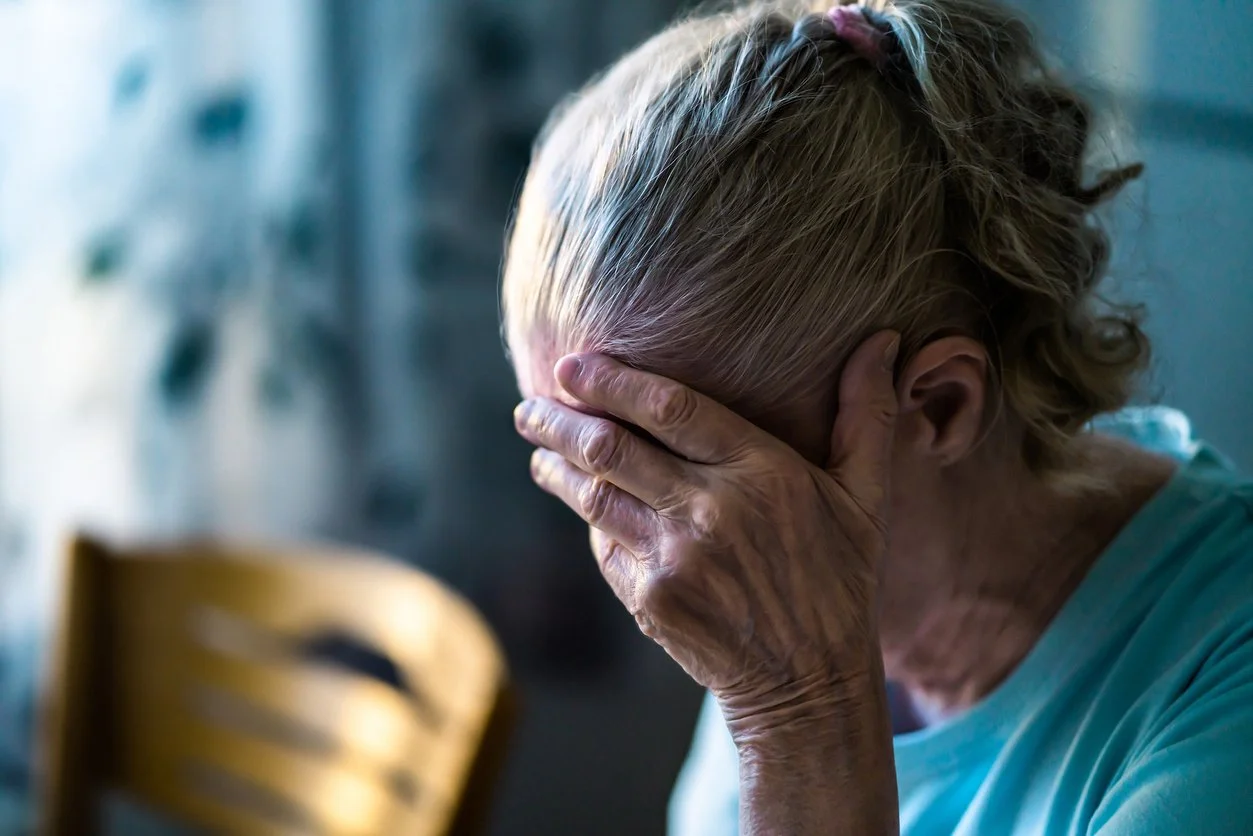 An elderly woman with gray hair sits at a table, holding her head with one hand, appearing distressed or upset.