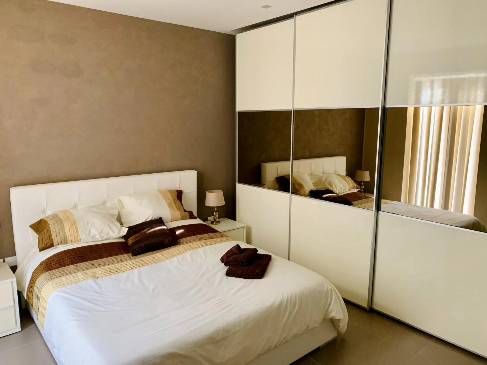 A modern bedroom with a large white bed, brown and beige striped bedding, and matching pillows. There are folded brown towels on the bed. A nightstand with a lamp is beside the bed, and there are built-in mirrored closet doors reflecting the room.