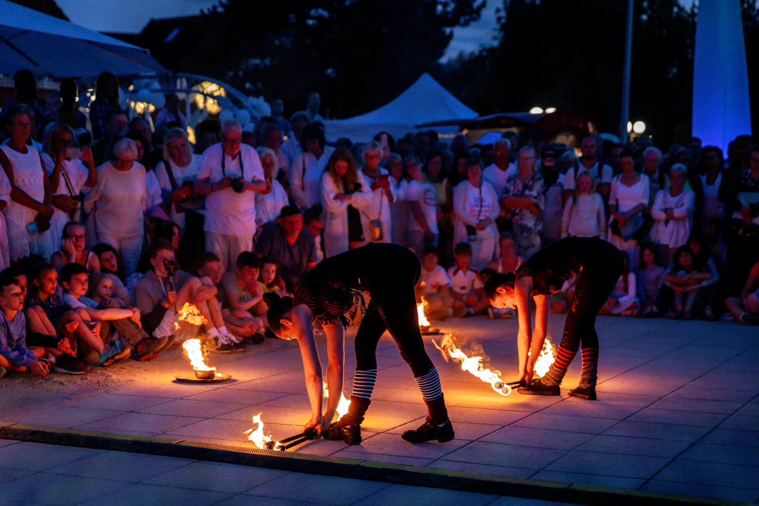 Two performers in black costumes and striped socks performing a fire routine on a stage in front of a large crowd at an outdoor event at dusk.