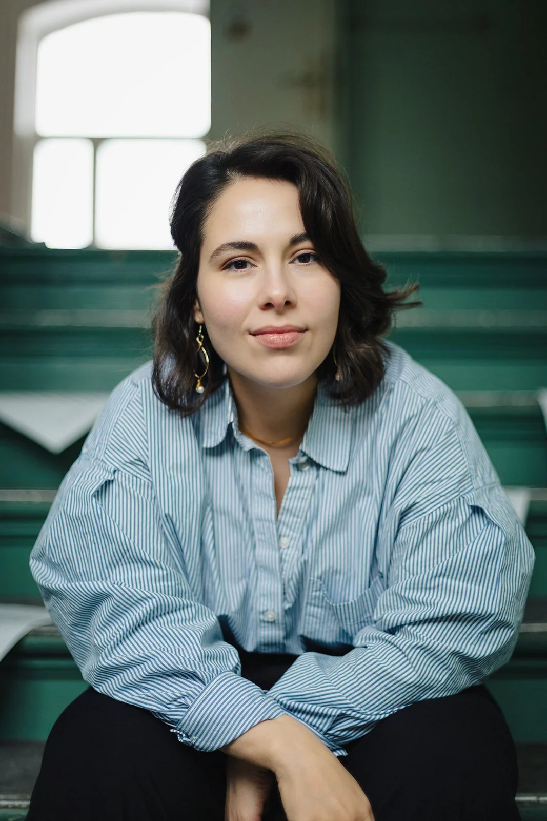 A young woman with dark, shoulder-length hair and earrings, sitting on green stadium stairs inside a building, wearing a striped button-up shirt.