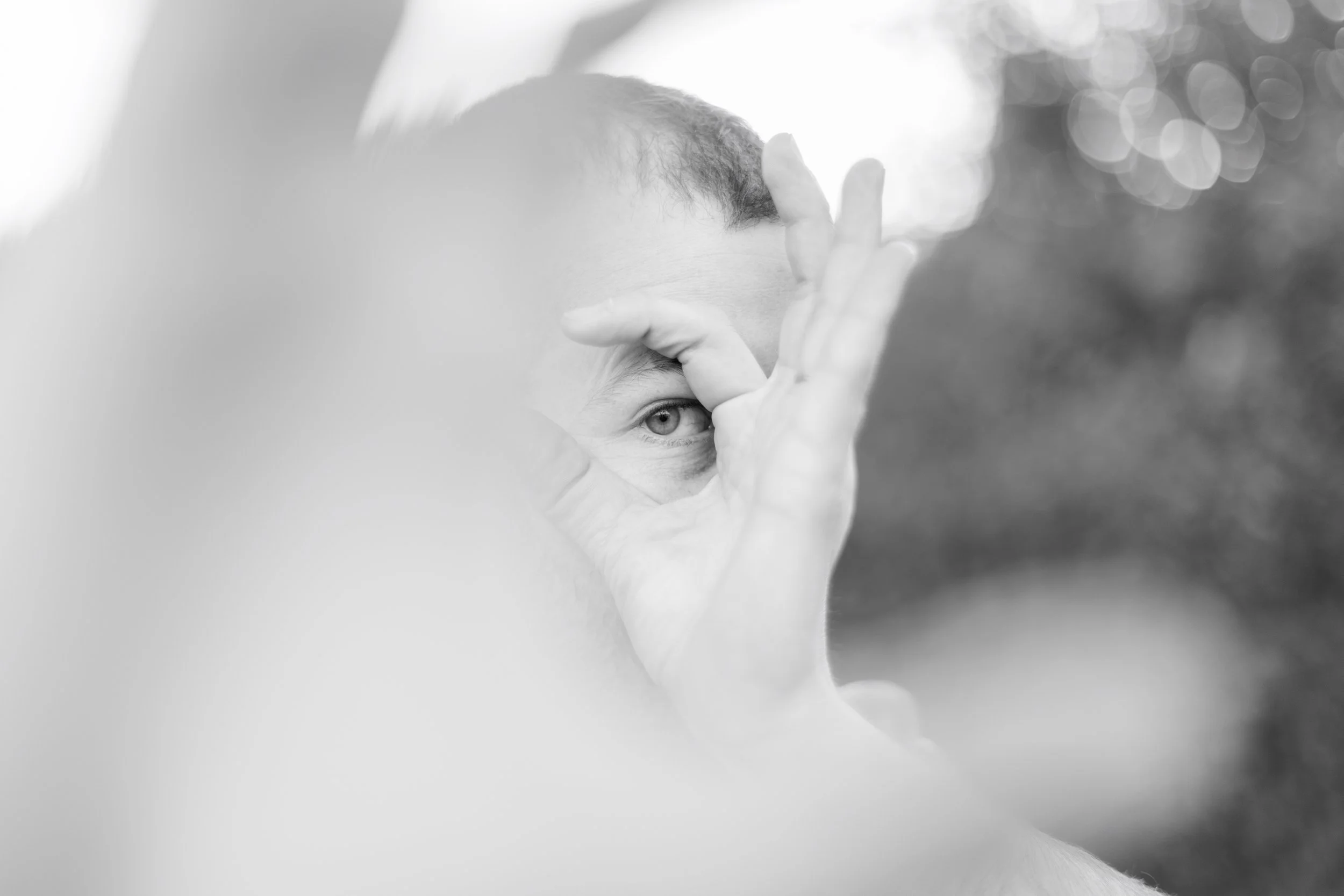 A person making a circle with their hand around one eye, looking through it, with a blurred outdoor background