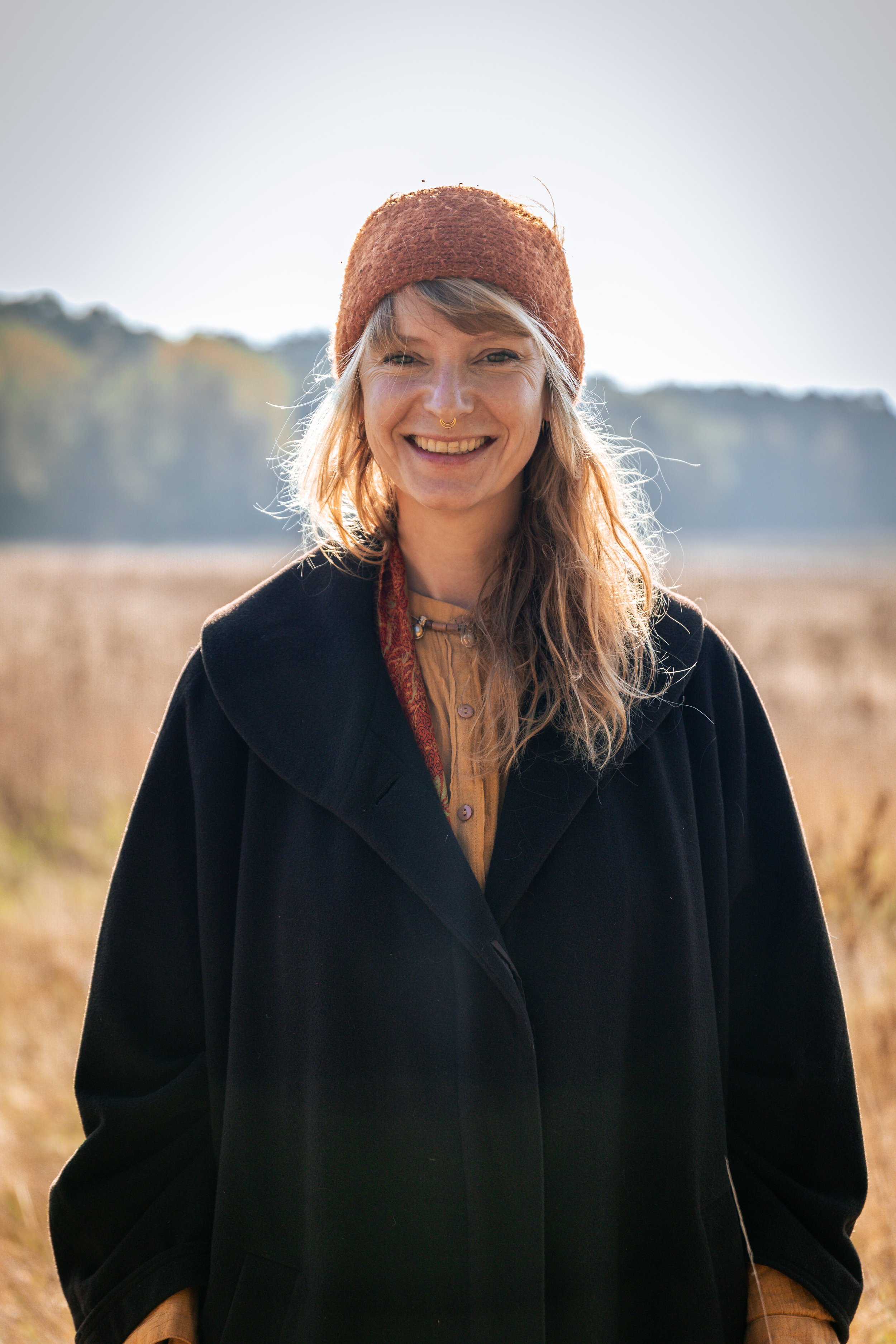 A woman is smiling outdoors in a field, wearing a brown beanie, black coat, and a mustard-colored shirt.