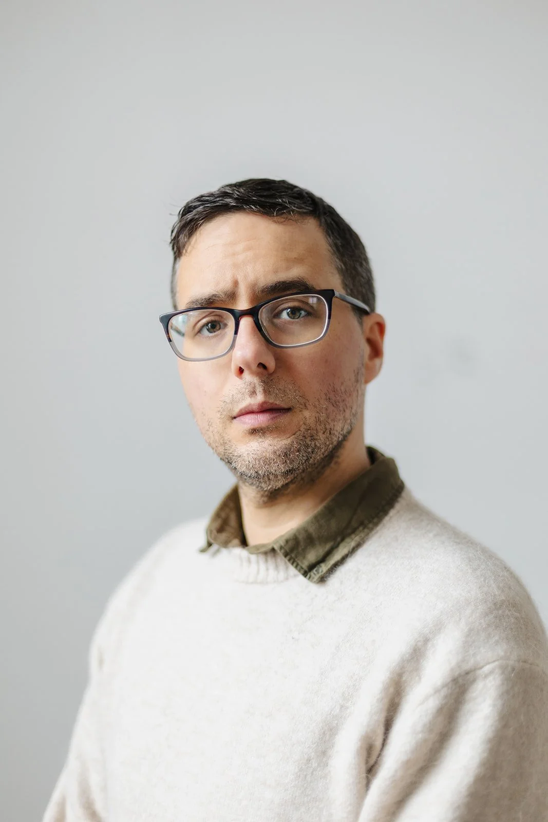 Portrait of a man wearing glasses, a beige sweater, and an olive green collared shirt, standing against a plain light gray background.