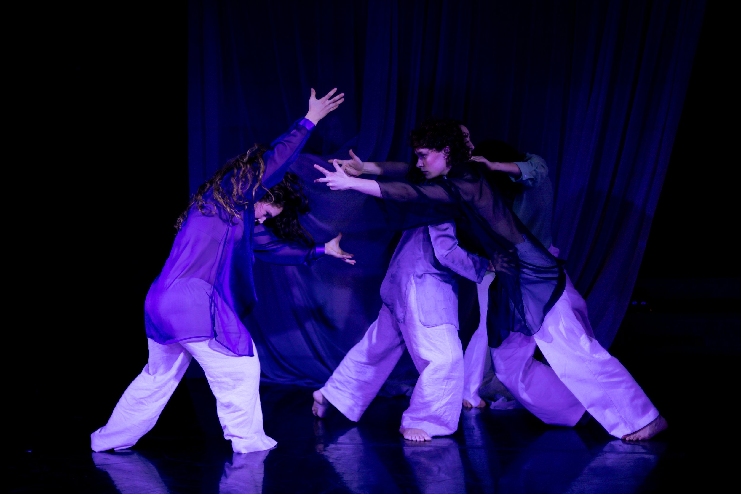 Dancer group performing on stage with purple lighting, wearing loose pants and sheer tops, against a dark backdrop.