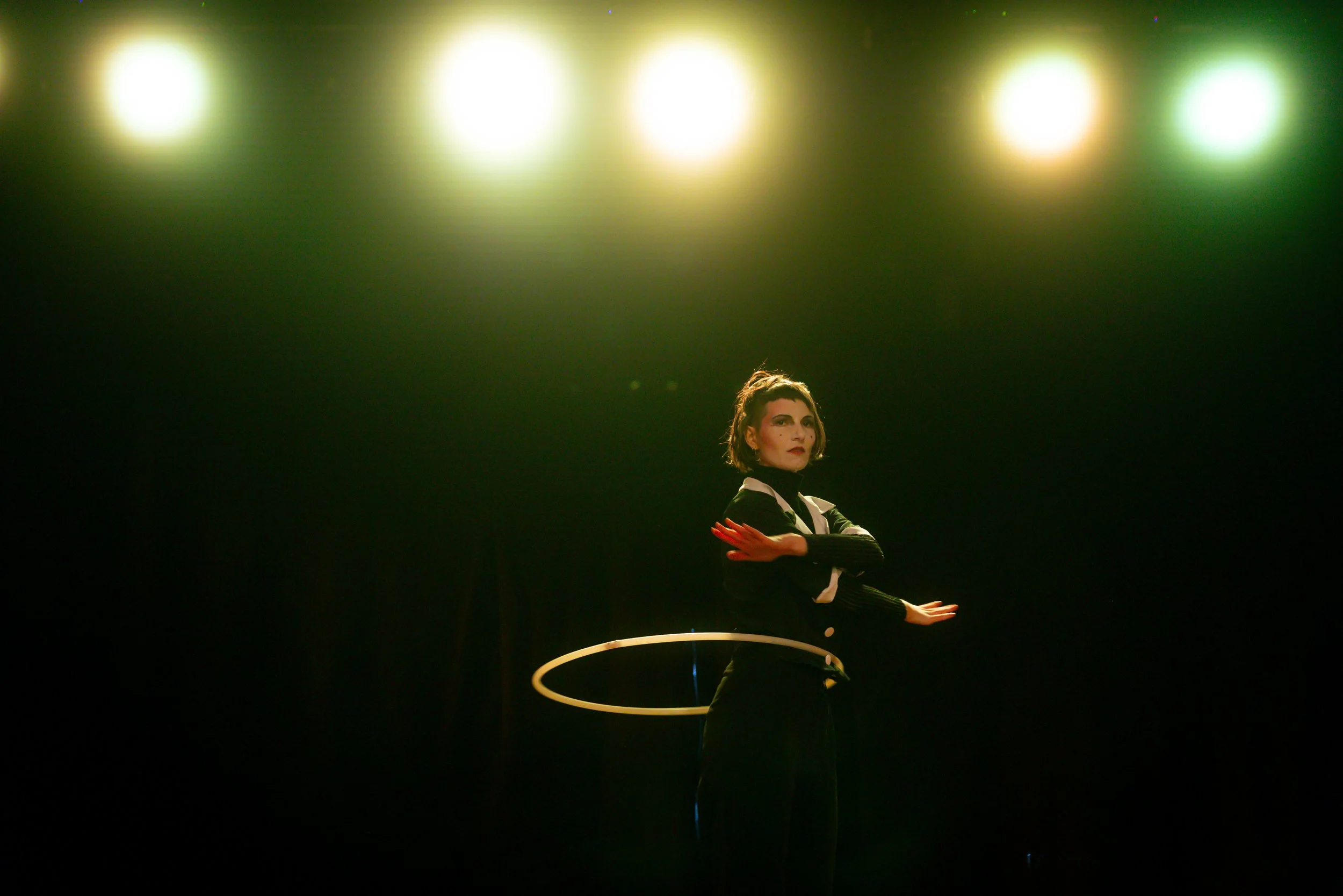 A woman standing on stage under bright yellow and green stage lights, wearing a black and white outfit and performing with a hula hoop.