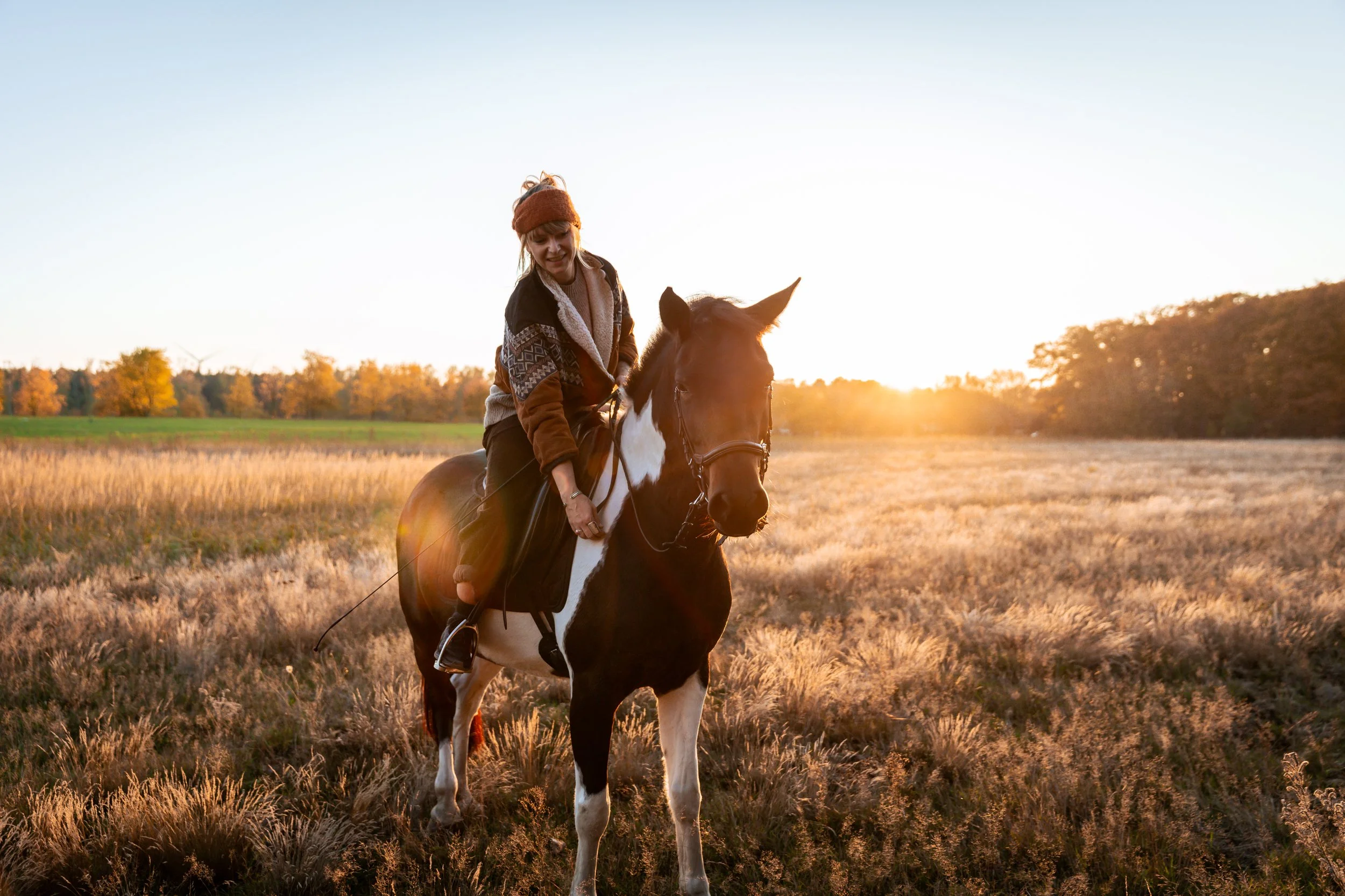 A woman riding a horse in a field at sunset, wearing a warm jacket and headband.
