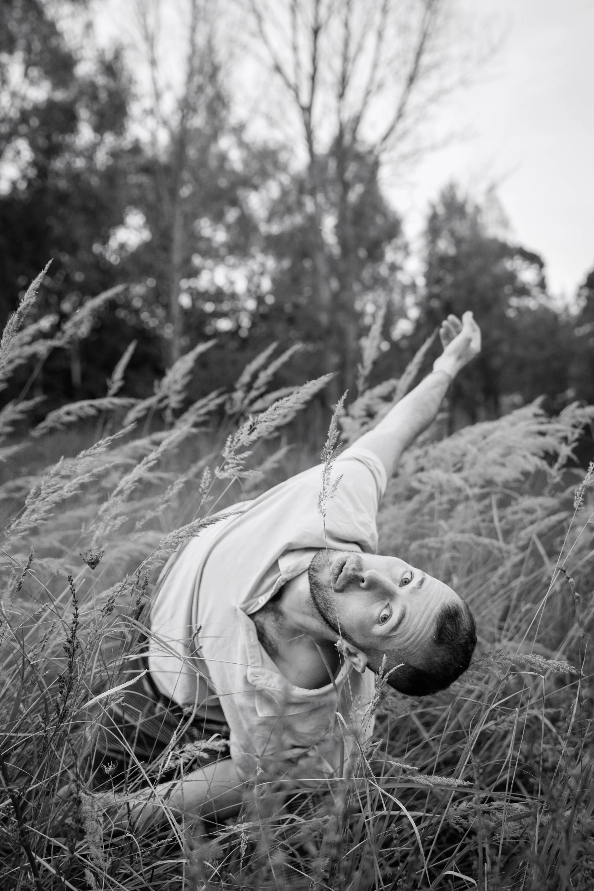 A man in a light-colored shirt crouching in tall grass, leaning to his right, with arms extended and looking at the camera, in a field with trees in the background, in black and white.