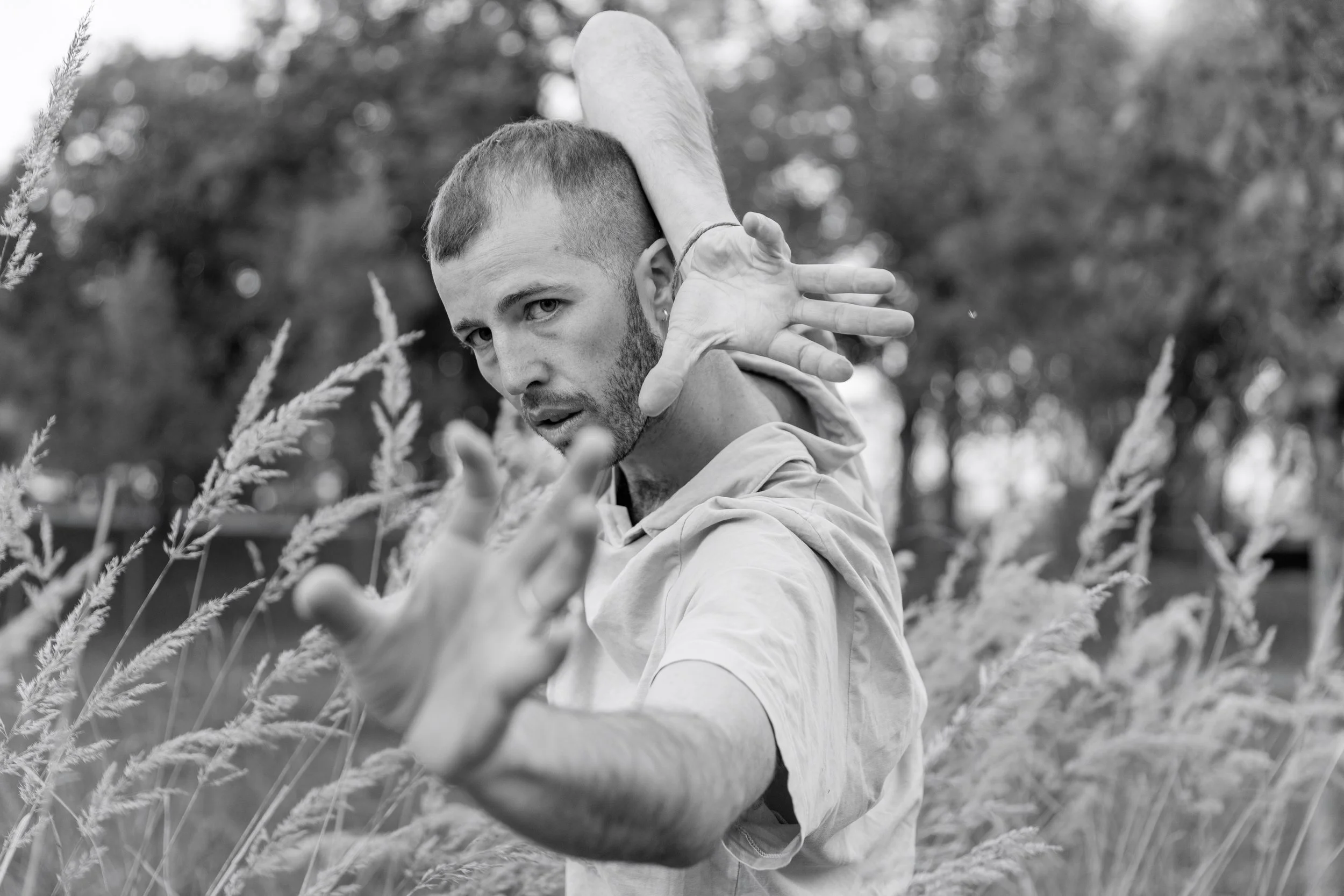 A man with short hair and a beard performing a dance or martial arts pose outdoors among tall grasses, with trees in the background.
