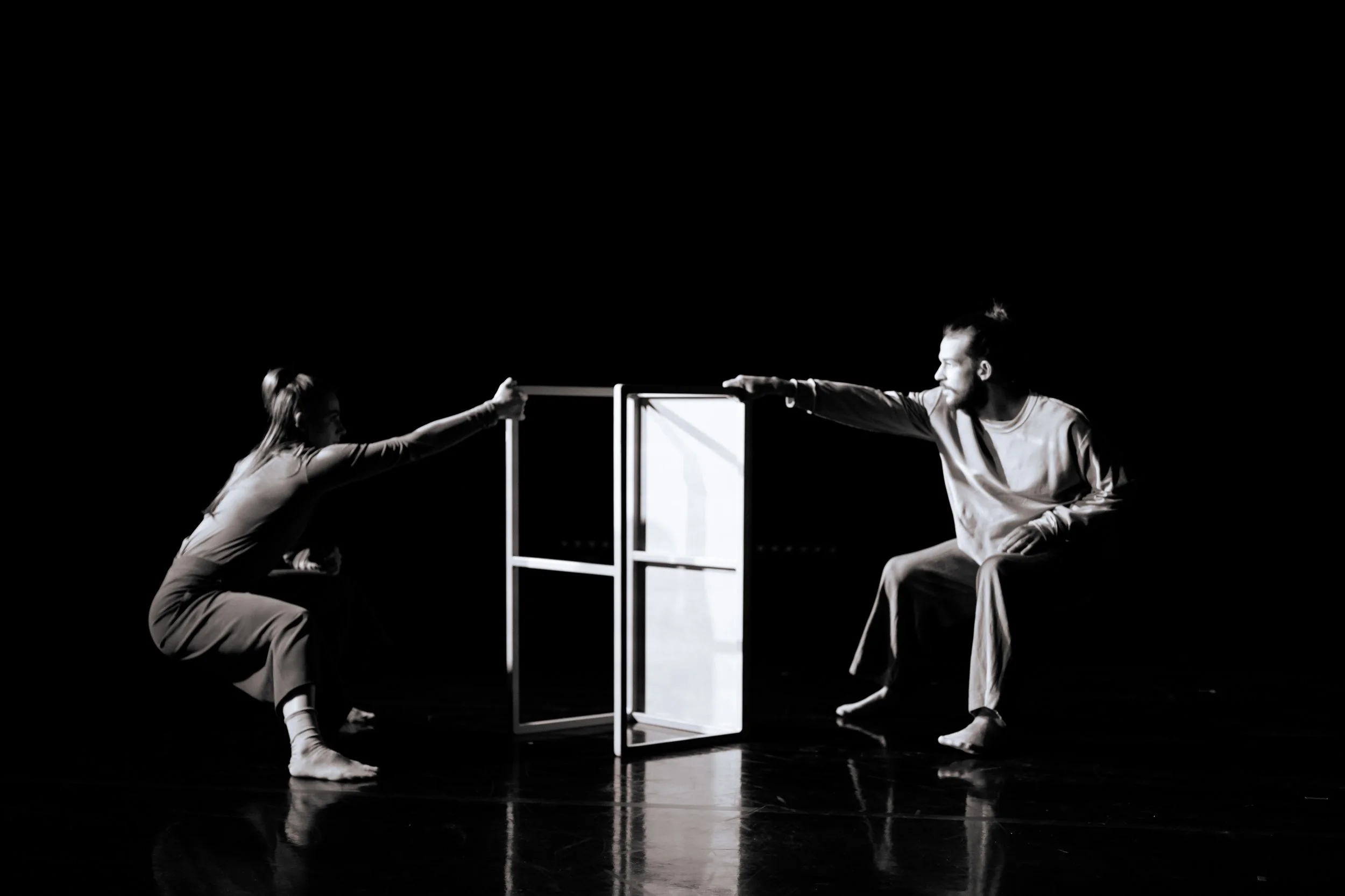 Two performers in casual clothing engaging in a choreographed dance or theater scene with a minimalistic set, including a frame and a door, on a dark stage.