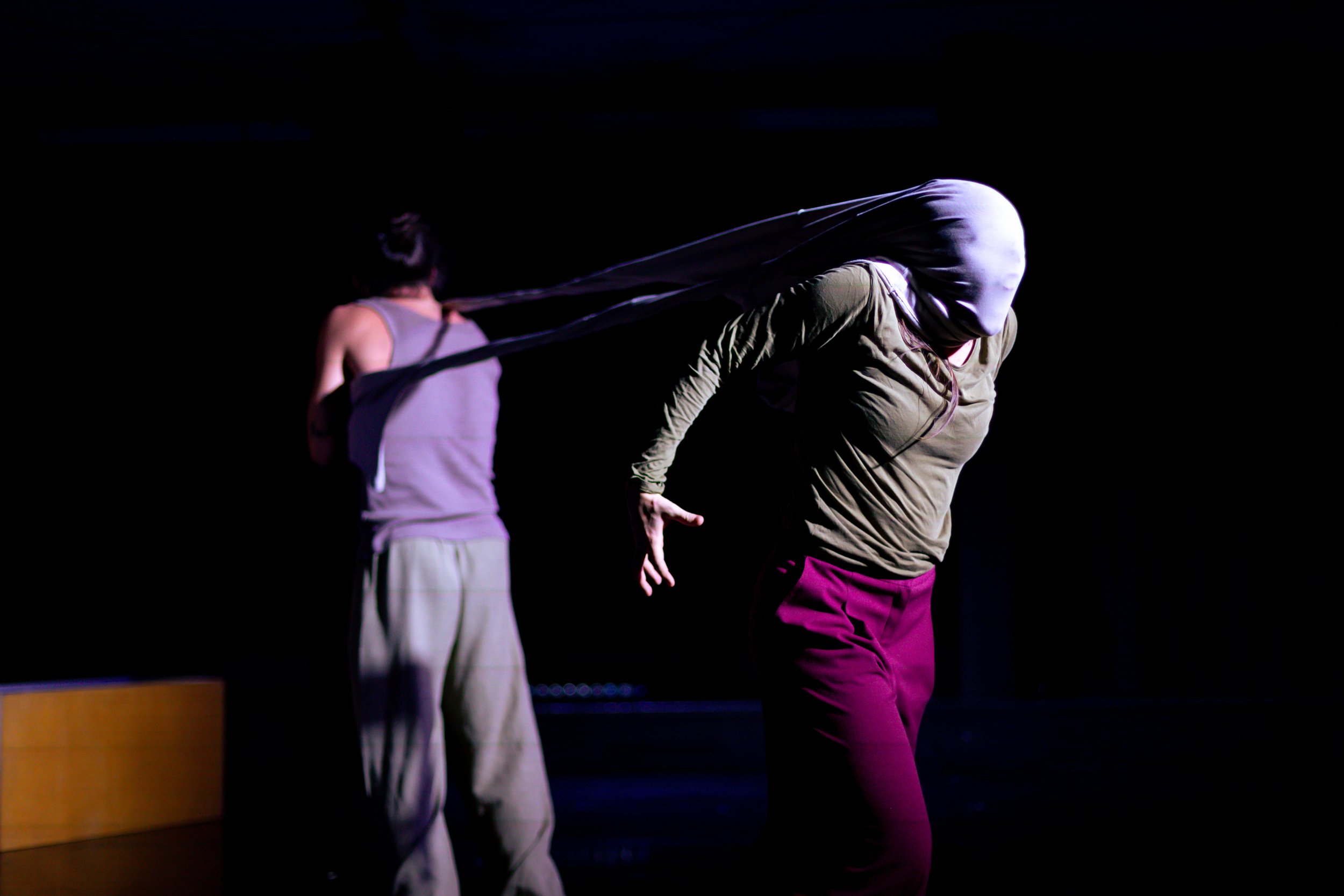 Two performers on stage, one with a white cloth over their face and head, reach out with her arm, while the other in the background has their back turned and is holding a long stretch of fabric or material between them in a dark, black background set