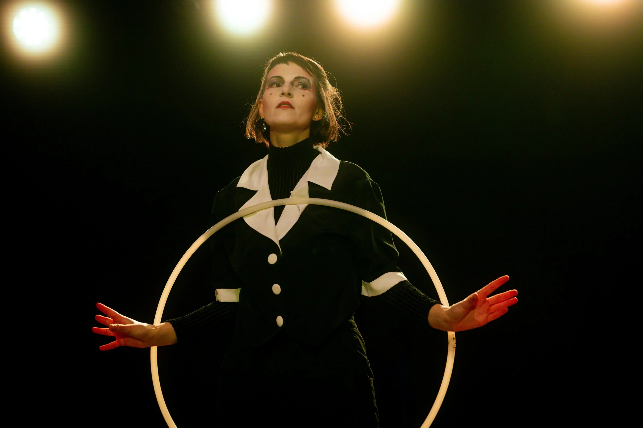 A woman dressed in a vintage black and white outfit is standing inside a glowing circle, with dramatic lighting highlighting her face and outfit.