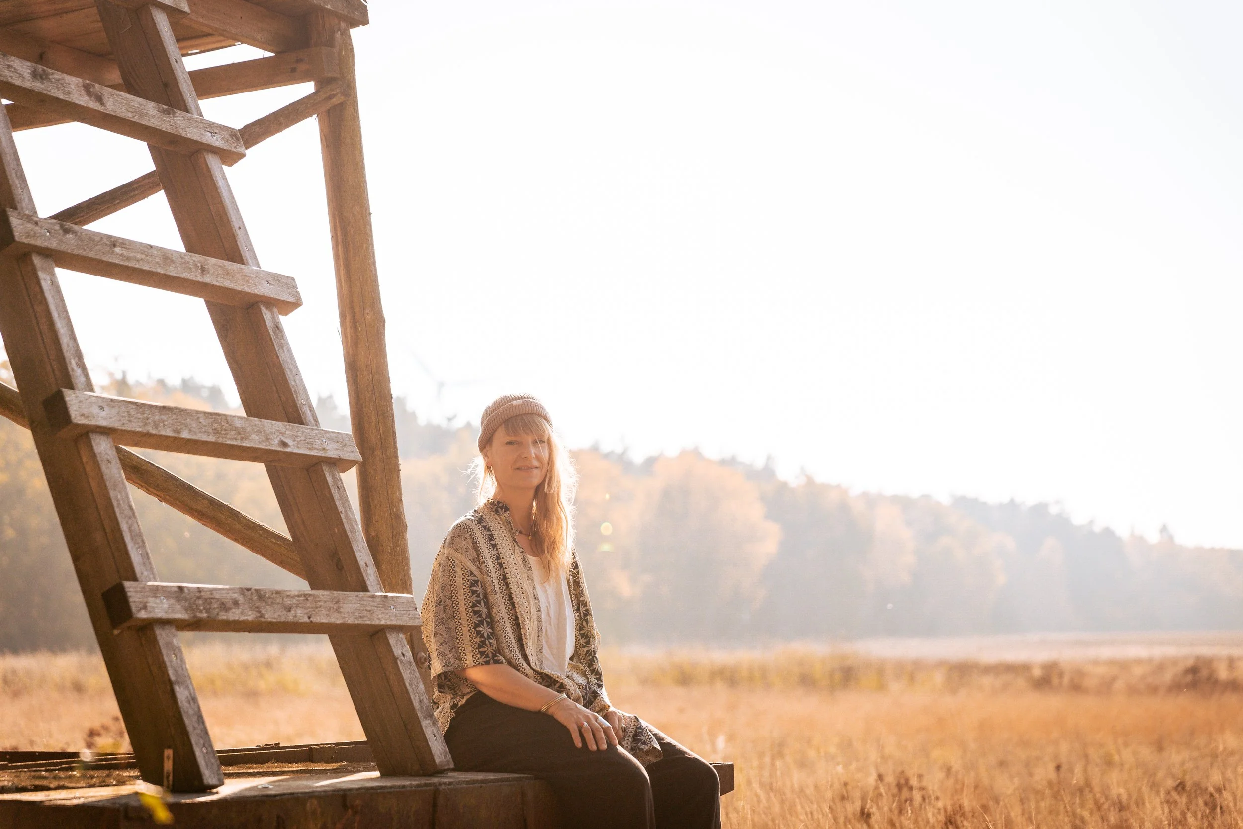 A woman sitting near a wooden windmill structure outdoors in a field during sunset, with trees in the background.