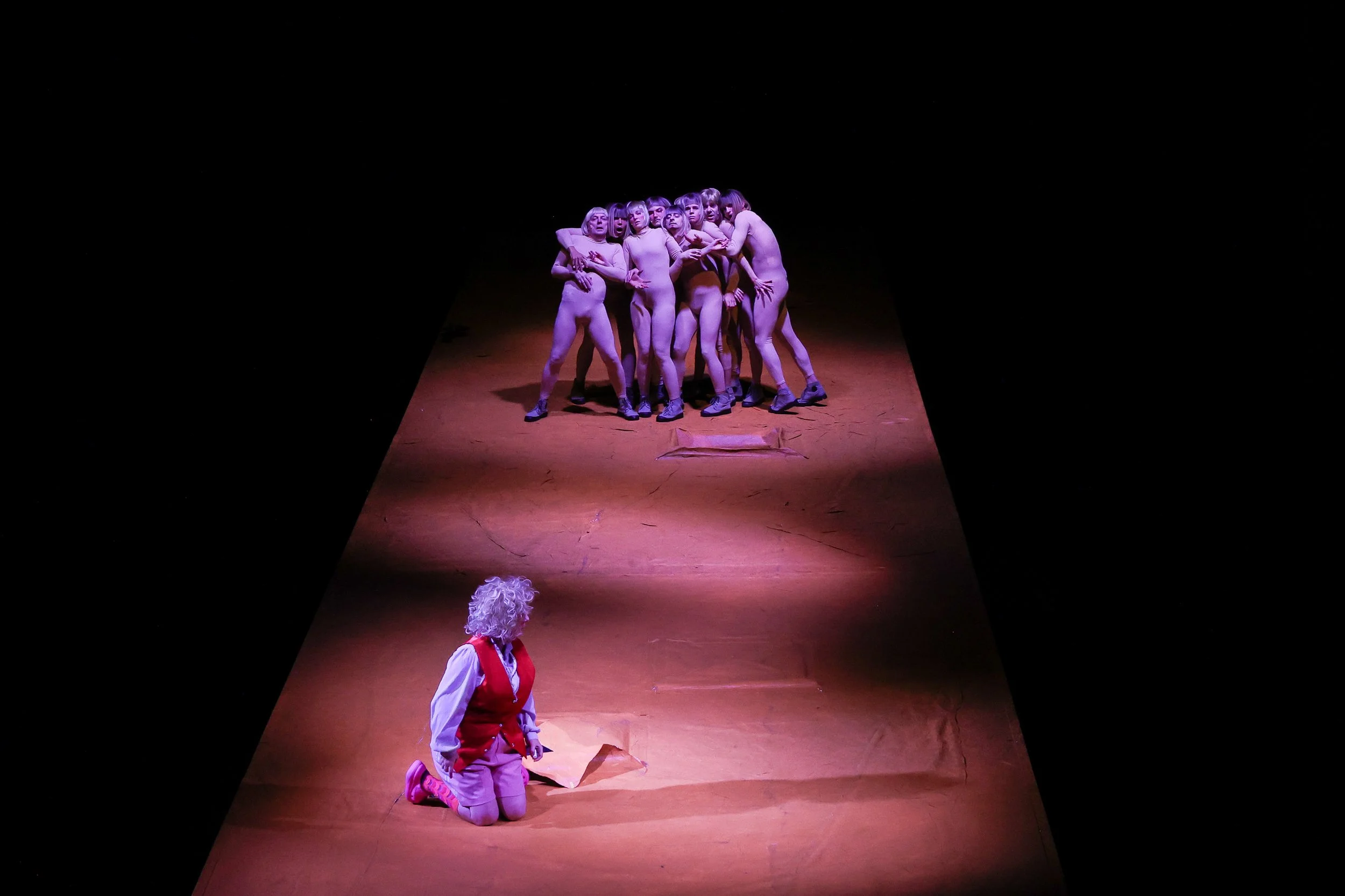 A theatrical stage scene with a person in a red vest and pink shoes kneeling on the floor, facing a group of performers with purple lighting, who are standing close together in a cluster.