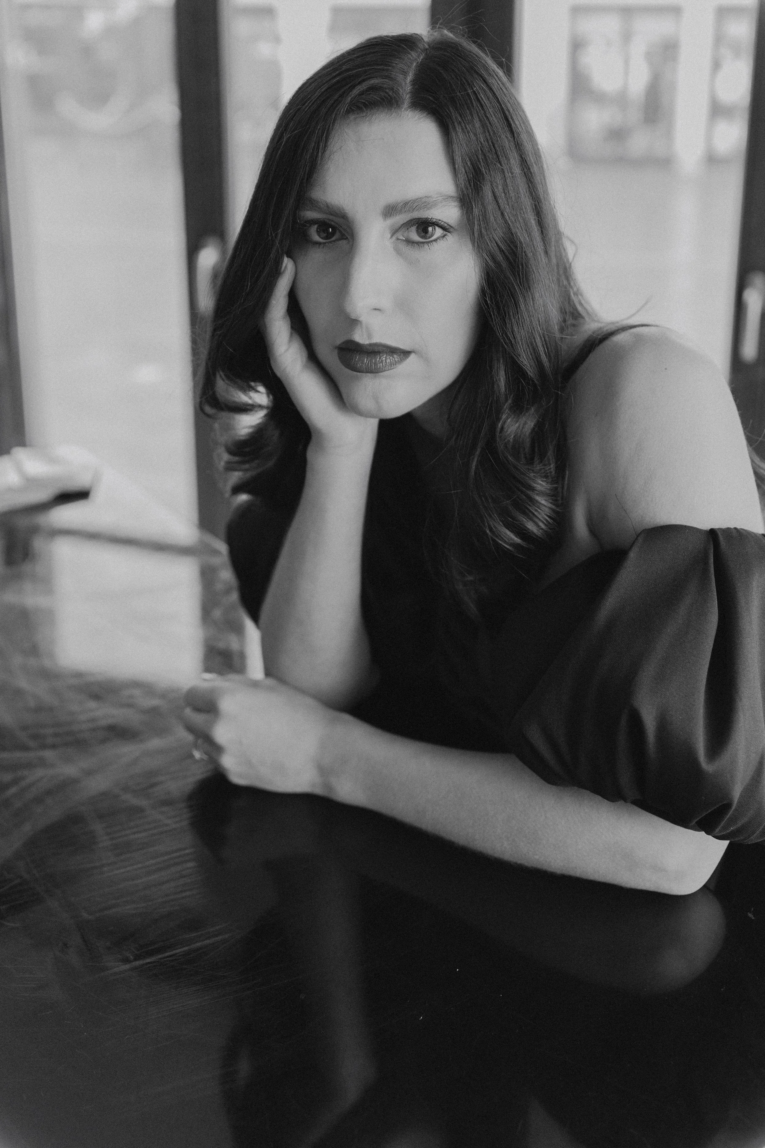 Black and white photo of a woman with long hair, resting her head on her hand, sitting at a table, looking directly at the camera.