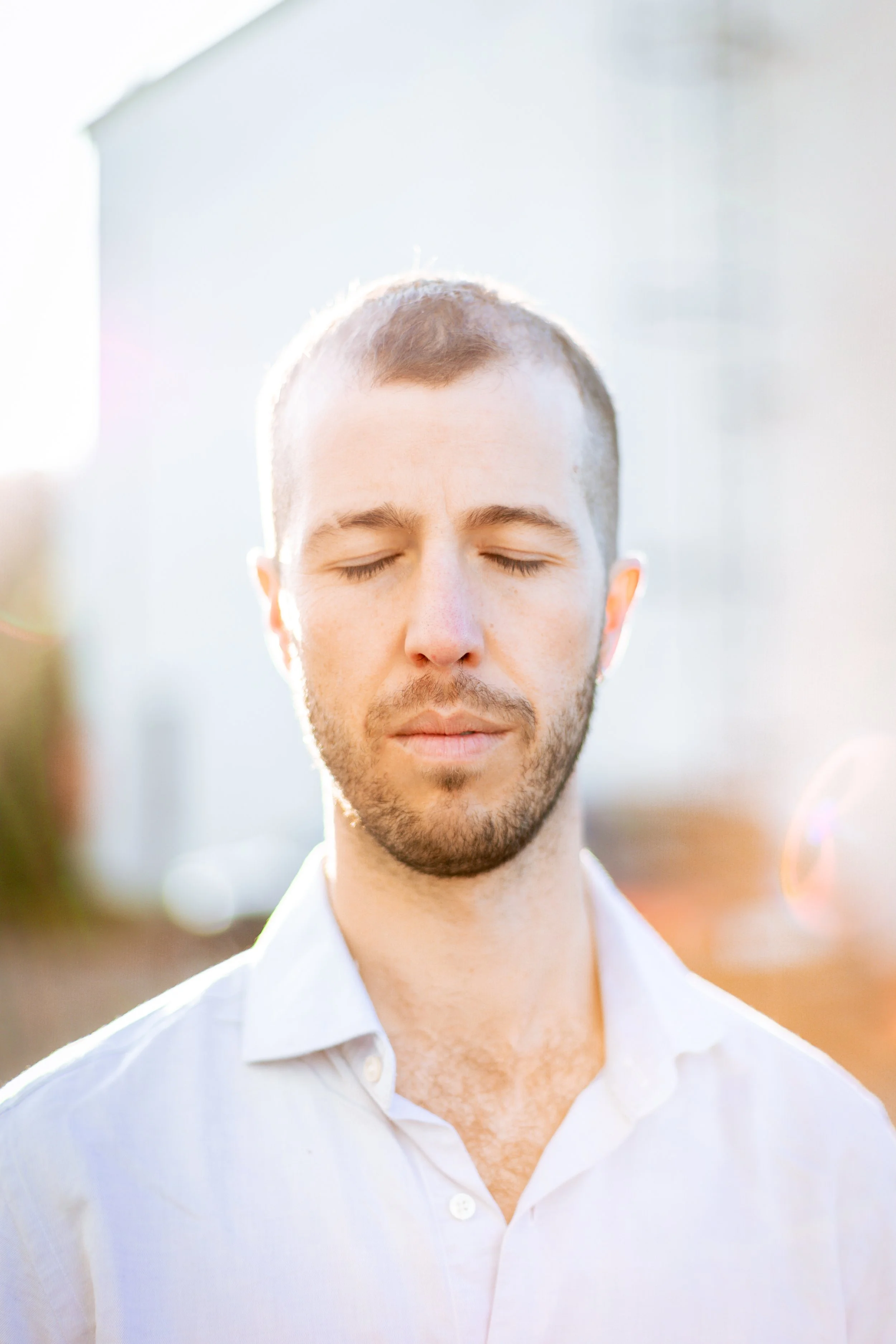 Close-up of a young man with short brown hair and a beard, wearing a white shirt, with eyes closed and a peaceful expression, outdoors during sunlight.