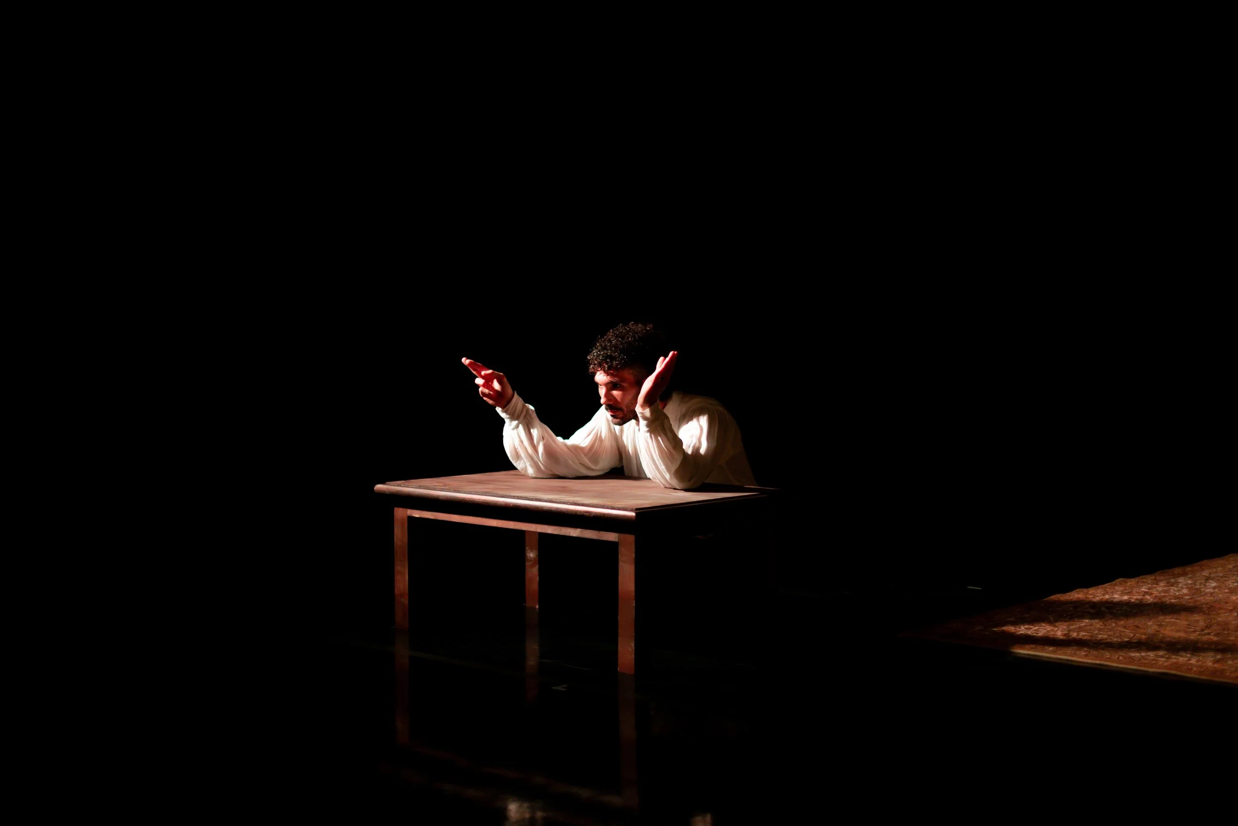 A man with curly hair and a beard, dressed in a white shirt, is sitting at a plain wooden table. He is making a gesture with both hands raised, with a surprised or expressive look on his face. The background is dark, highlighting the man and the tabl