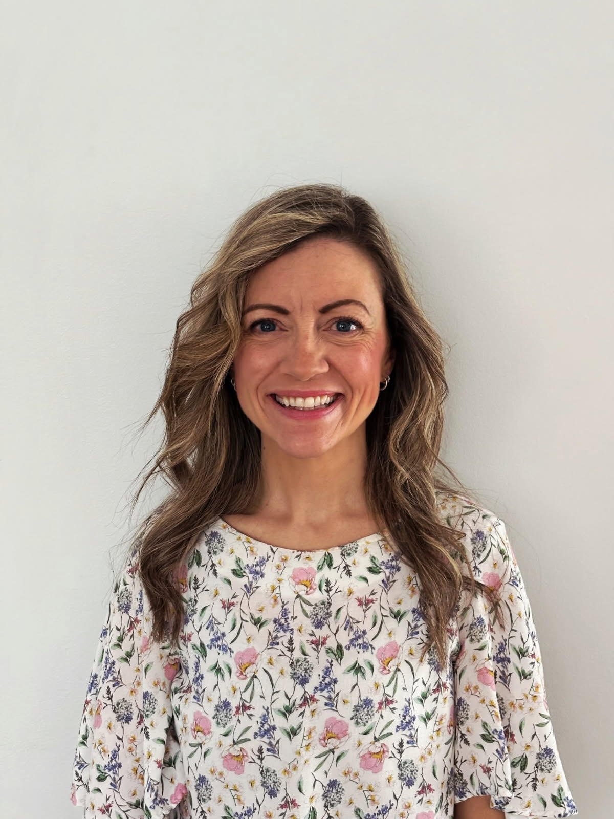 A woman with wavy light brown hair, blue eyes, and a broad smile, wearing a white floral blouse, standing against a plain white wall.