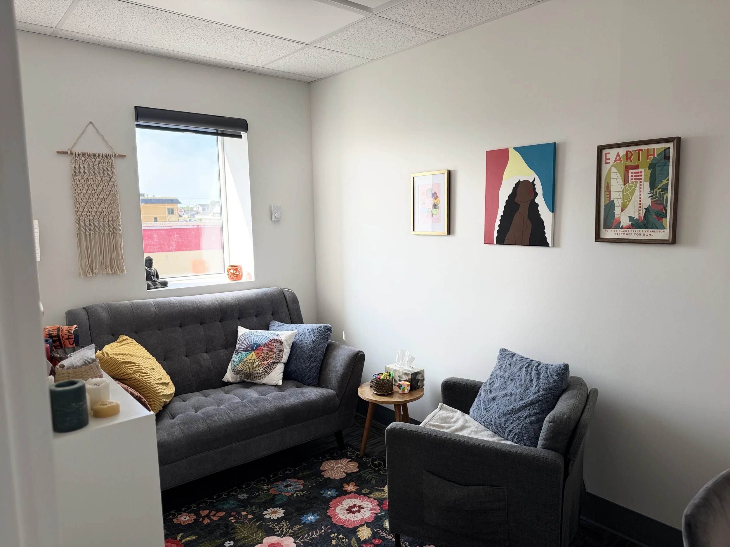 A cozy living room with a gray sofa and single armchair, decorated with colorful cushions. There is a small wooden side table with tissues and small objects, and a black floral rug. The wall behind the sofa has three framed artworks, including one wi