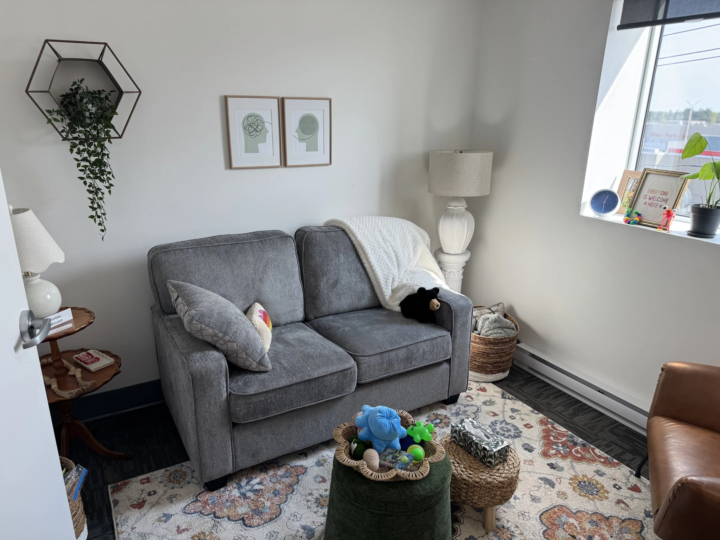 A cozy living room with a gray sofa, a white quilt draped over the arm, a teddy bear on the armrest, and a patterned cushion. There is a side table with a lamp and a phone, a lamp on a white stand beside the sofa, and a colorful rug on the floor. A b
