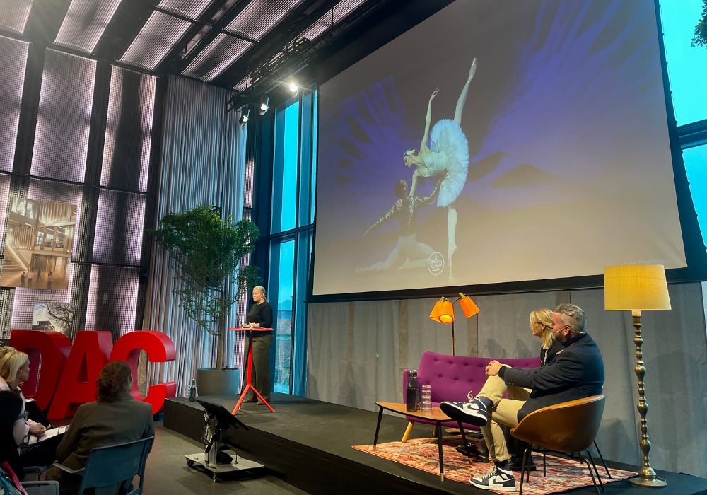 Nina Degn, Head of CX, The Royal Danish Theater speaking at a large black stage, in front of a big screen with an image of a ballet dancer.