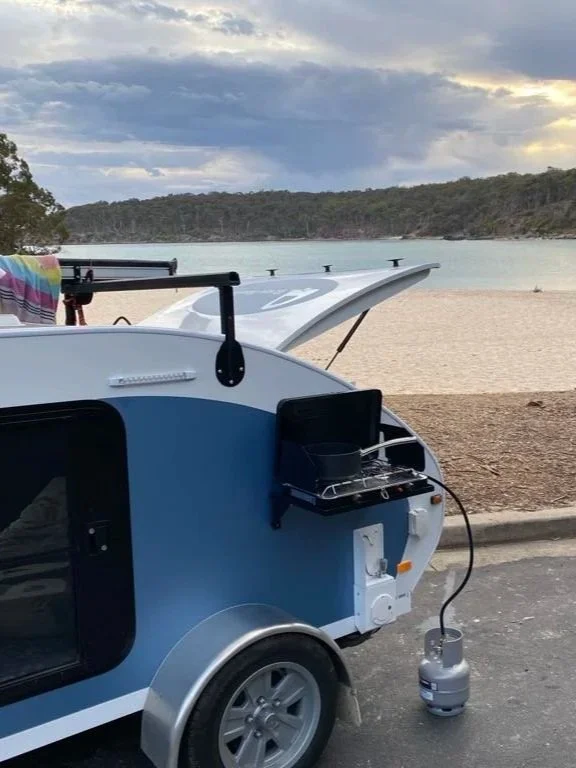 A small camper trailer with a gas stove and a propane tank parked near a beach with sand and water, hills in the background, cloudy sky overhead.