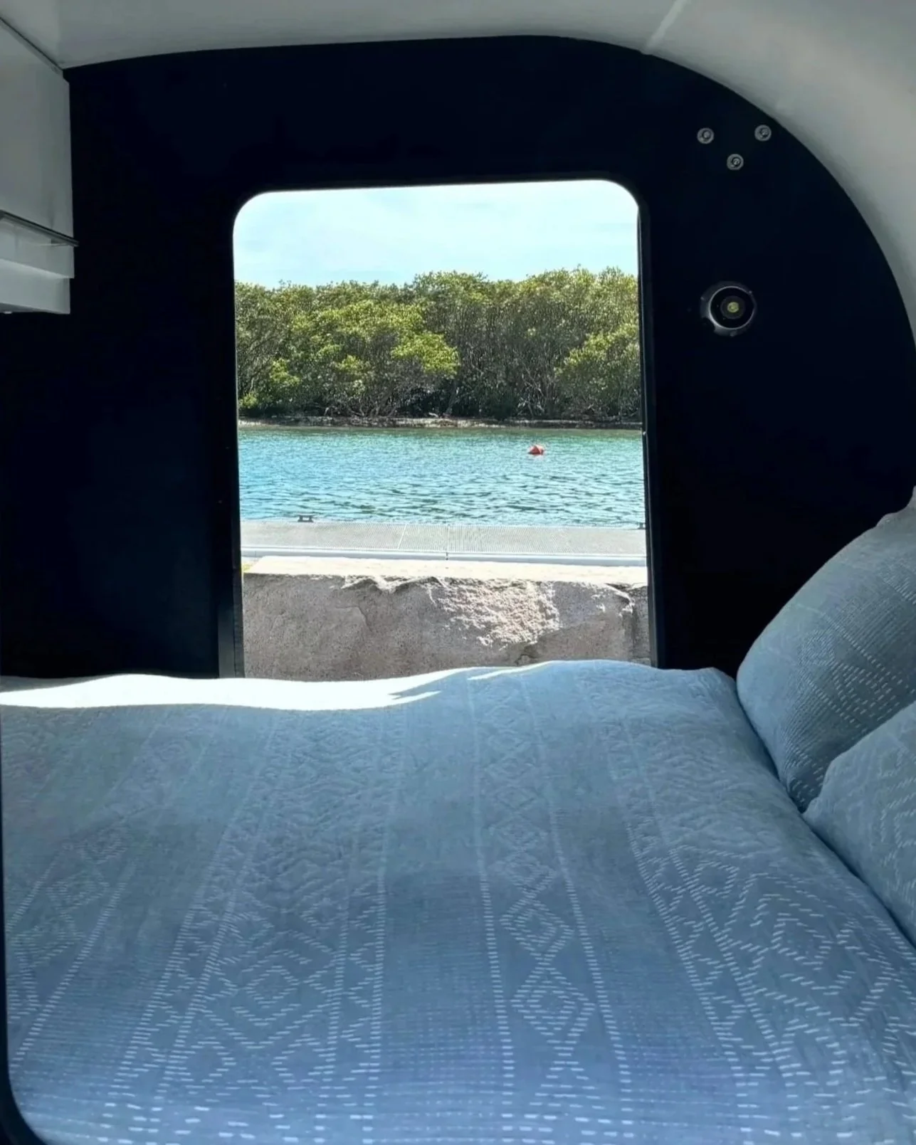 View of water and lush green trees outside a boat window, with a bed and white bedding inside the boat.