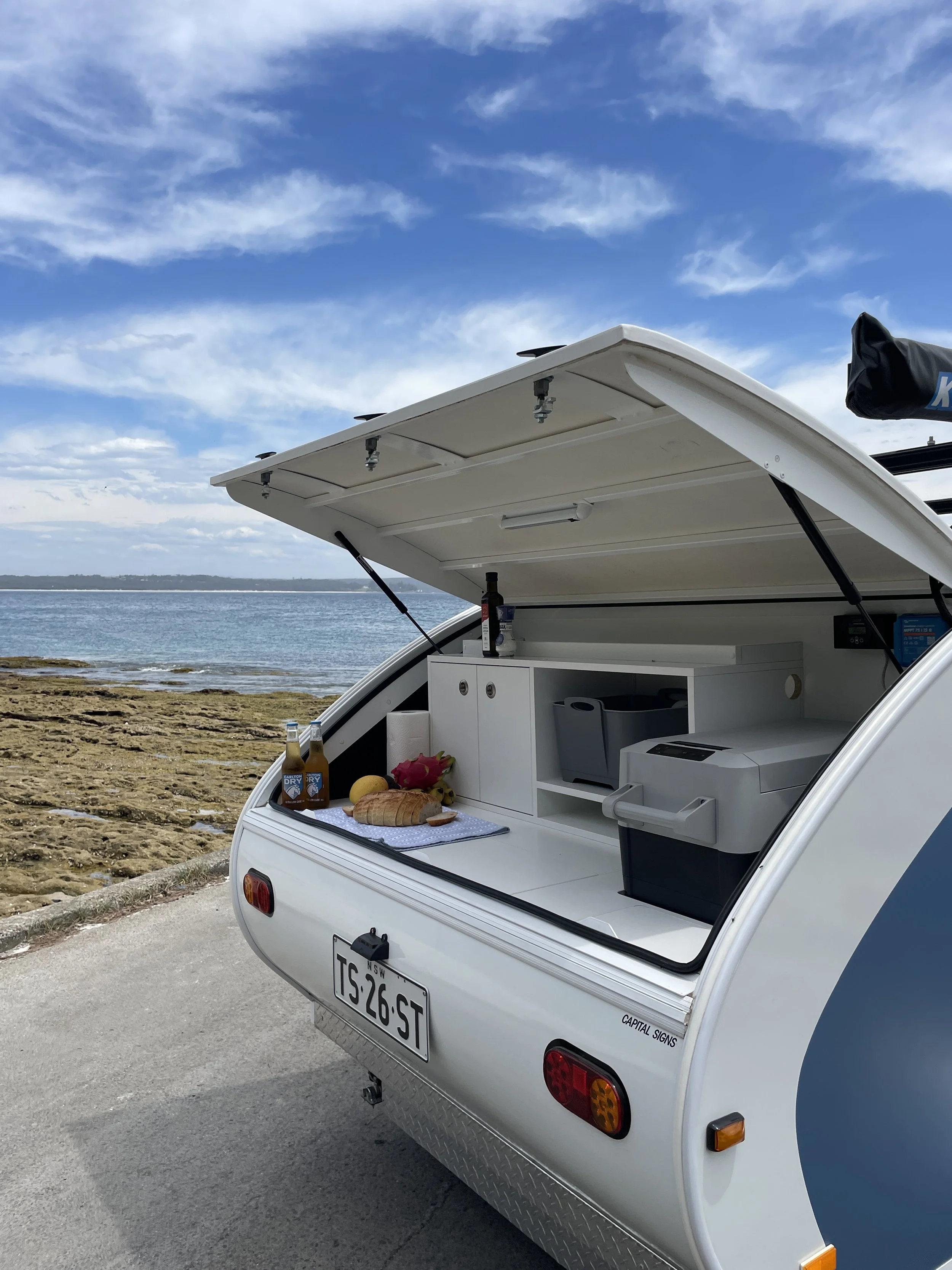 Back of a white camper trailer with its hatch open, showing a picnic setup with drinks, bread, fruit, and a small cooler, near the ocean with rocky shoreline and blue sky.