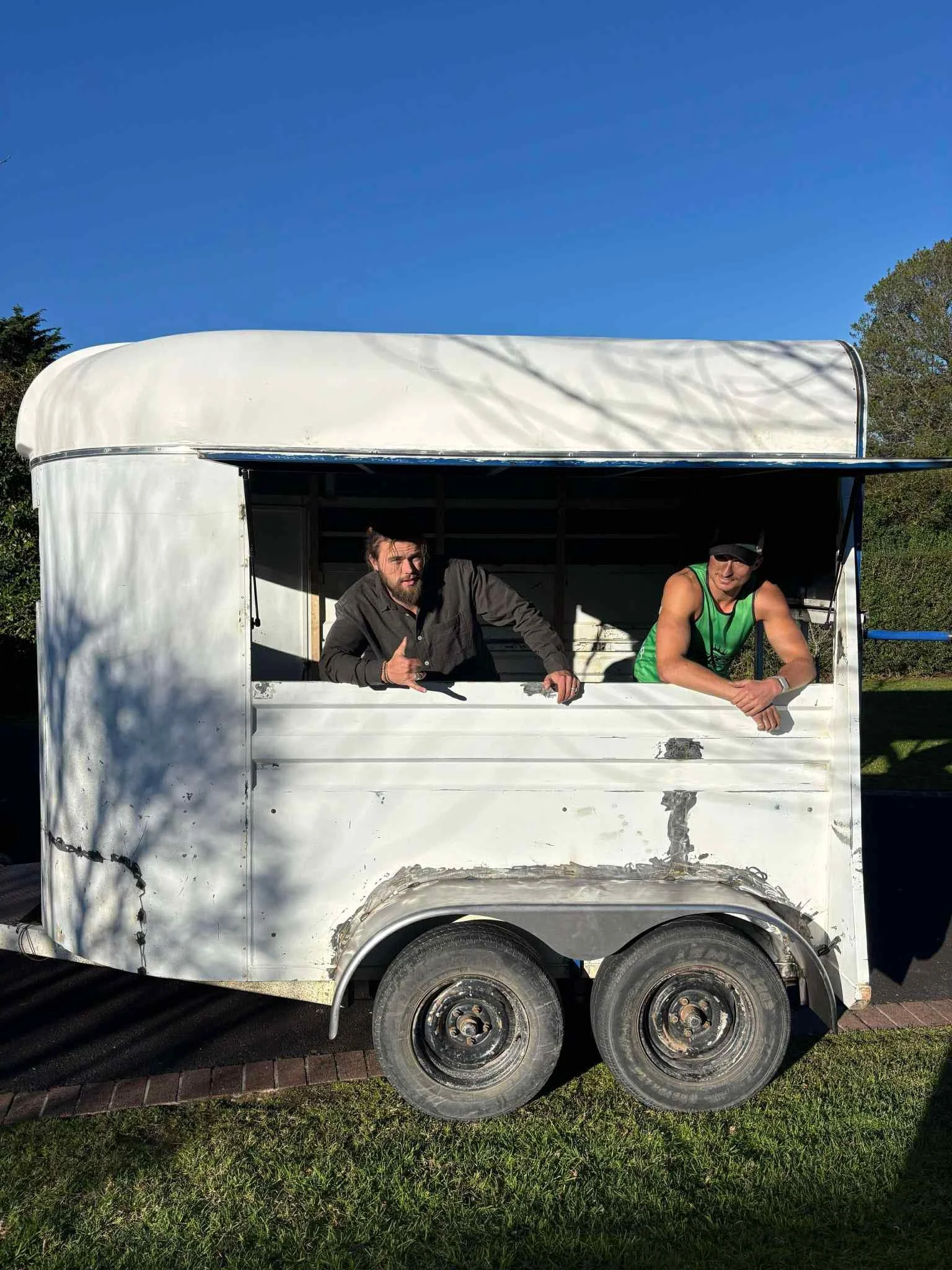 Two men leaning on a white trailer with two wheels, parked on a grassy area, with trees in the background and a clear blue sky.