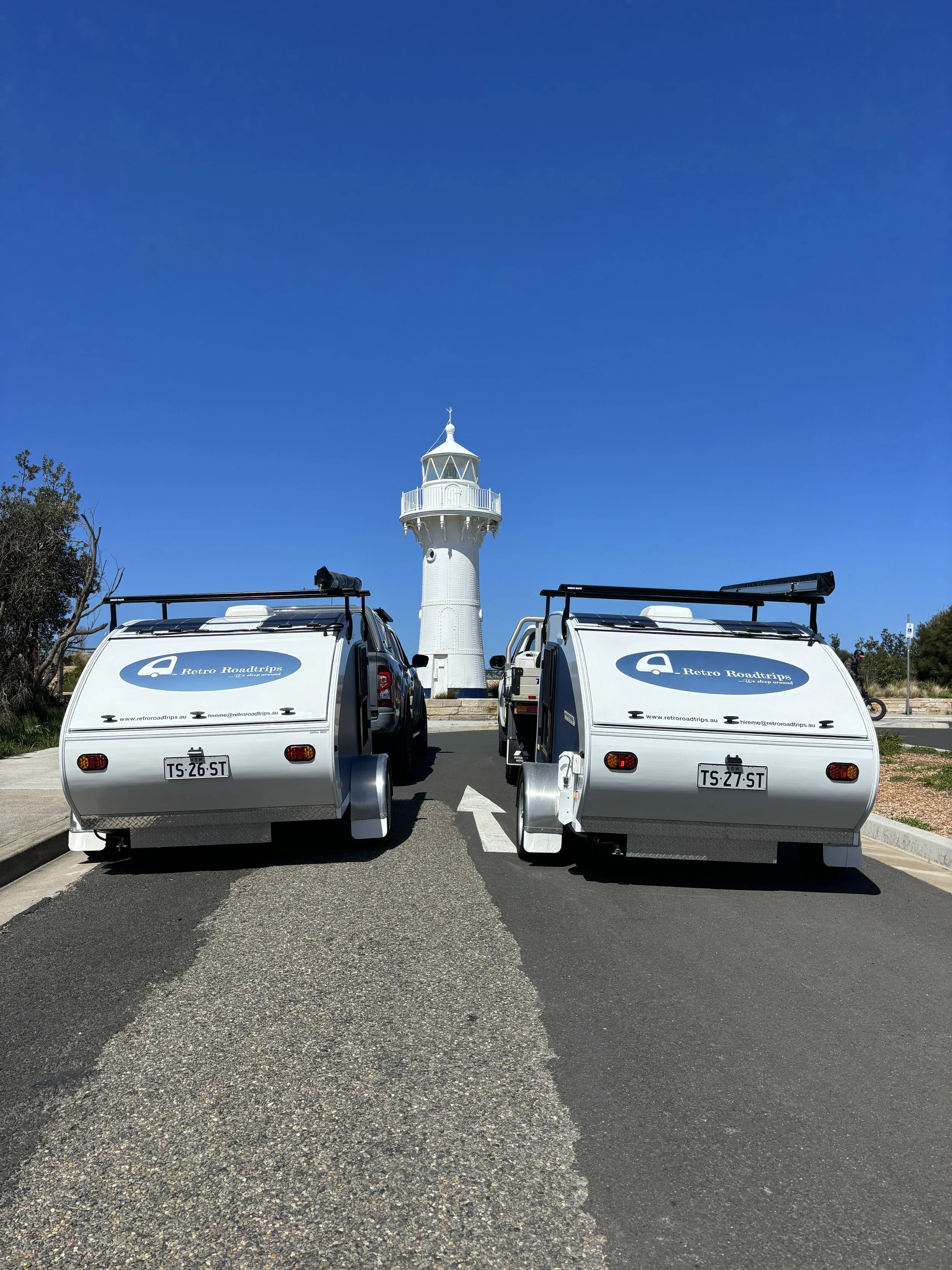 Two white trailers with 'Retro Roadtrips' logos on a paved road, parked in front of a white lighthouse under a clear blue sky.