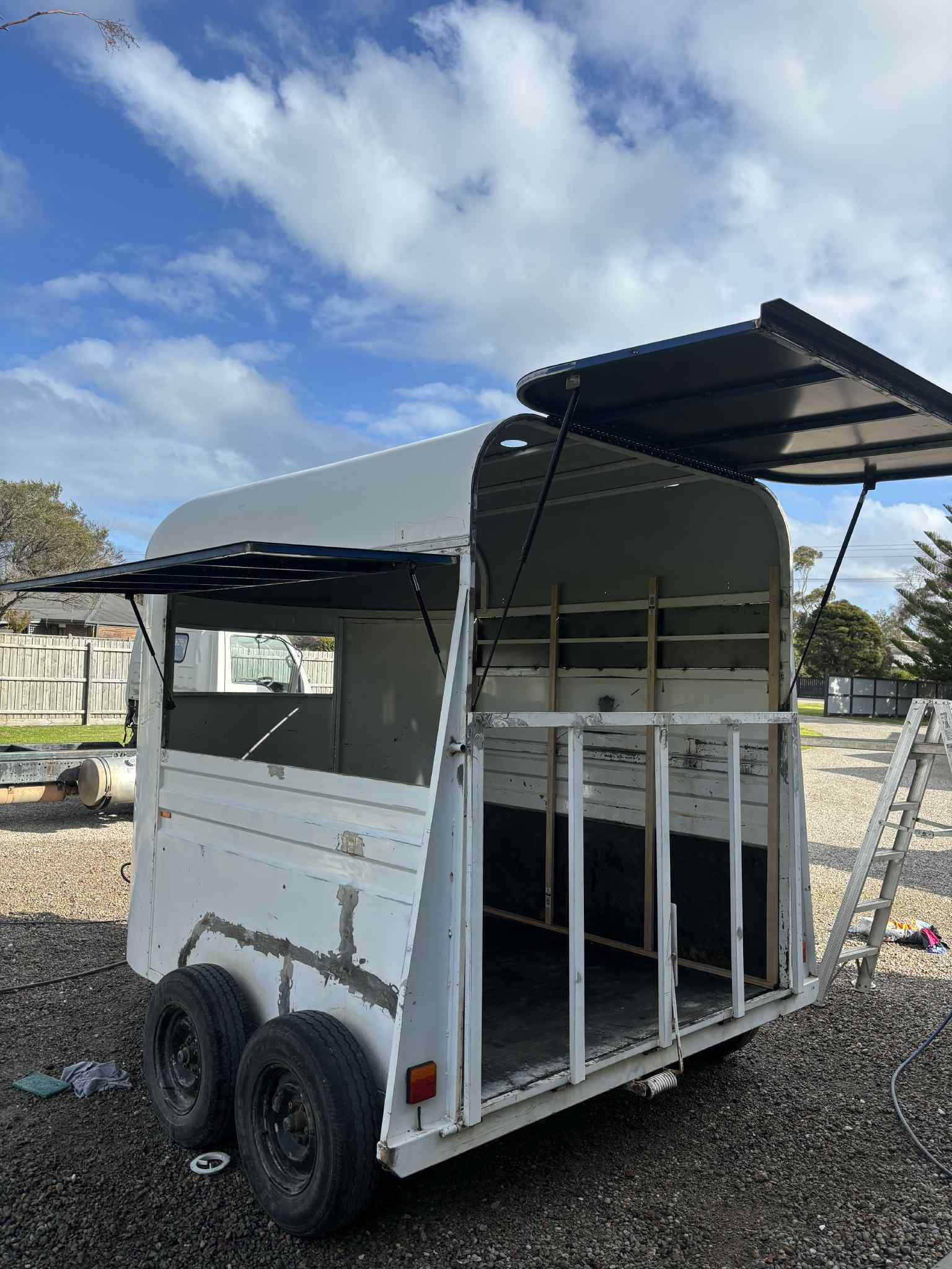 A white food truck or trailer with open side panels and a small ladder, parked on gravel with trees and a fence in the background under a partly cloudy sky.