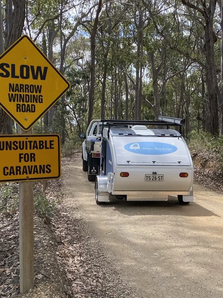 A narrow dirt road through a wooded area with a yellow sign reading "Slow Narrow Winding Road" and another sign saying "Unsuitable for Caravans." A small caravan is parked on the road behind a pickup truck.