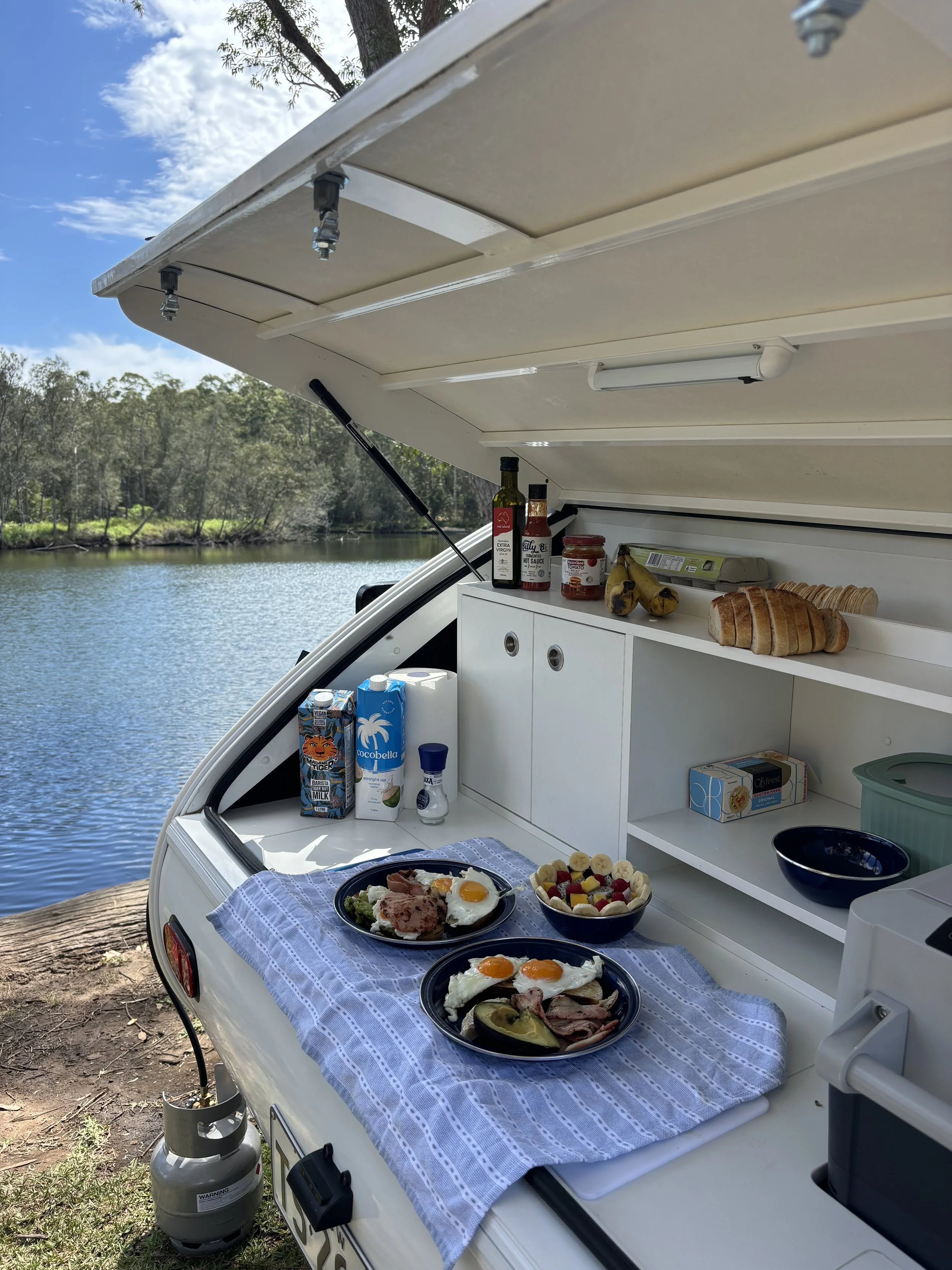 Breakfast spread on a boat served outdoors by a lake, with eggs, bacon, and other breakfast foods on plates, and milk, juice, bread, and condiments on the boat's storage area.
