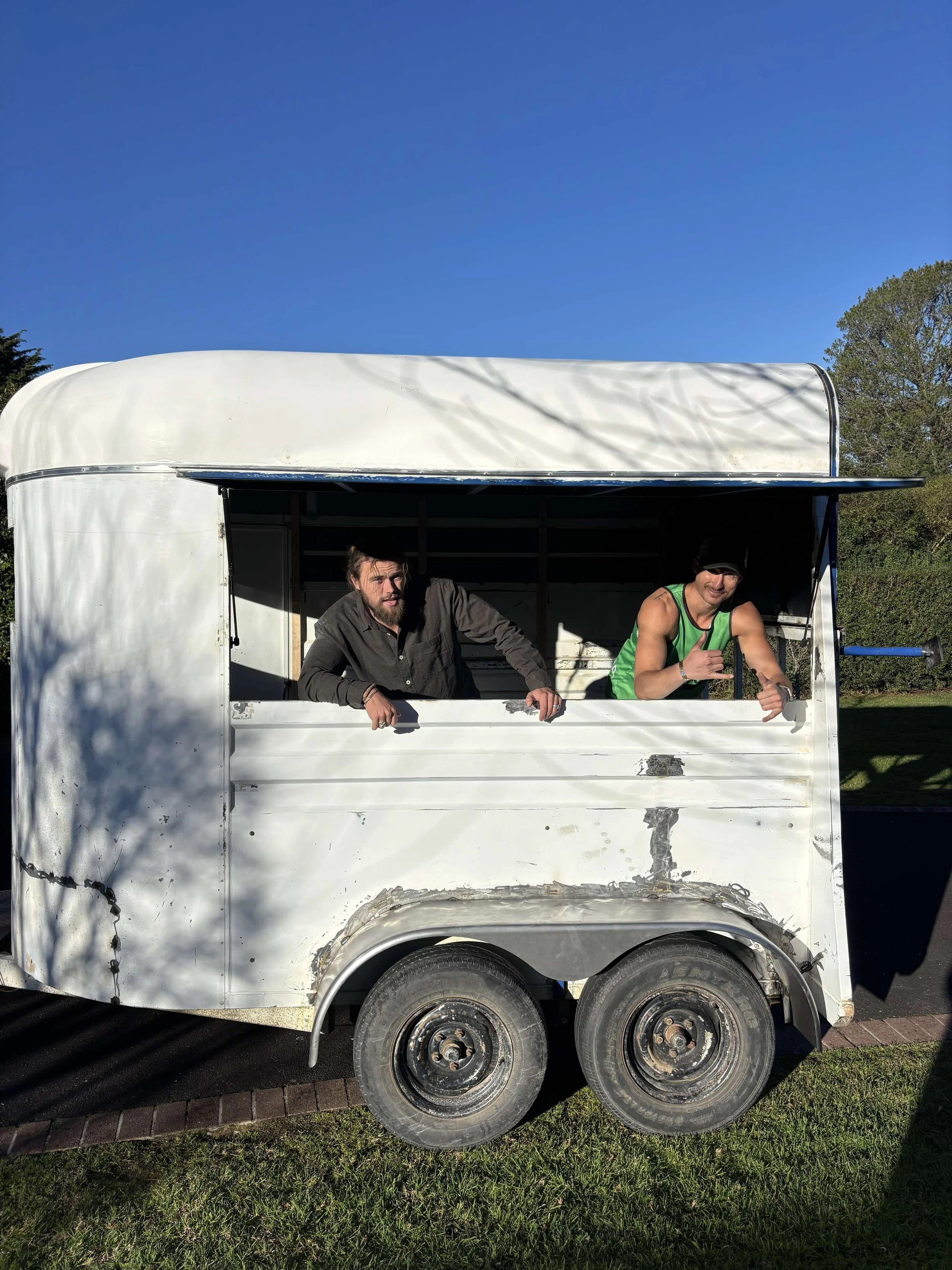 Two men leaning out of a white food truck with an open window, parked outdoors on a sunny day.