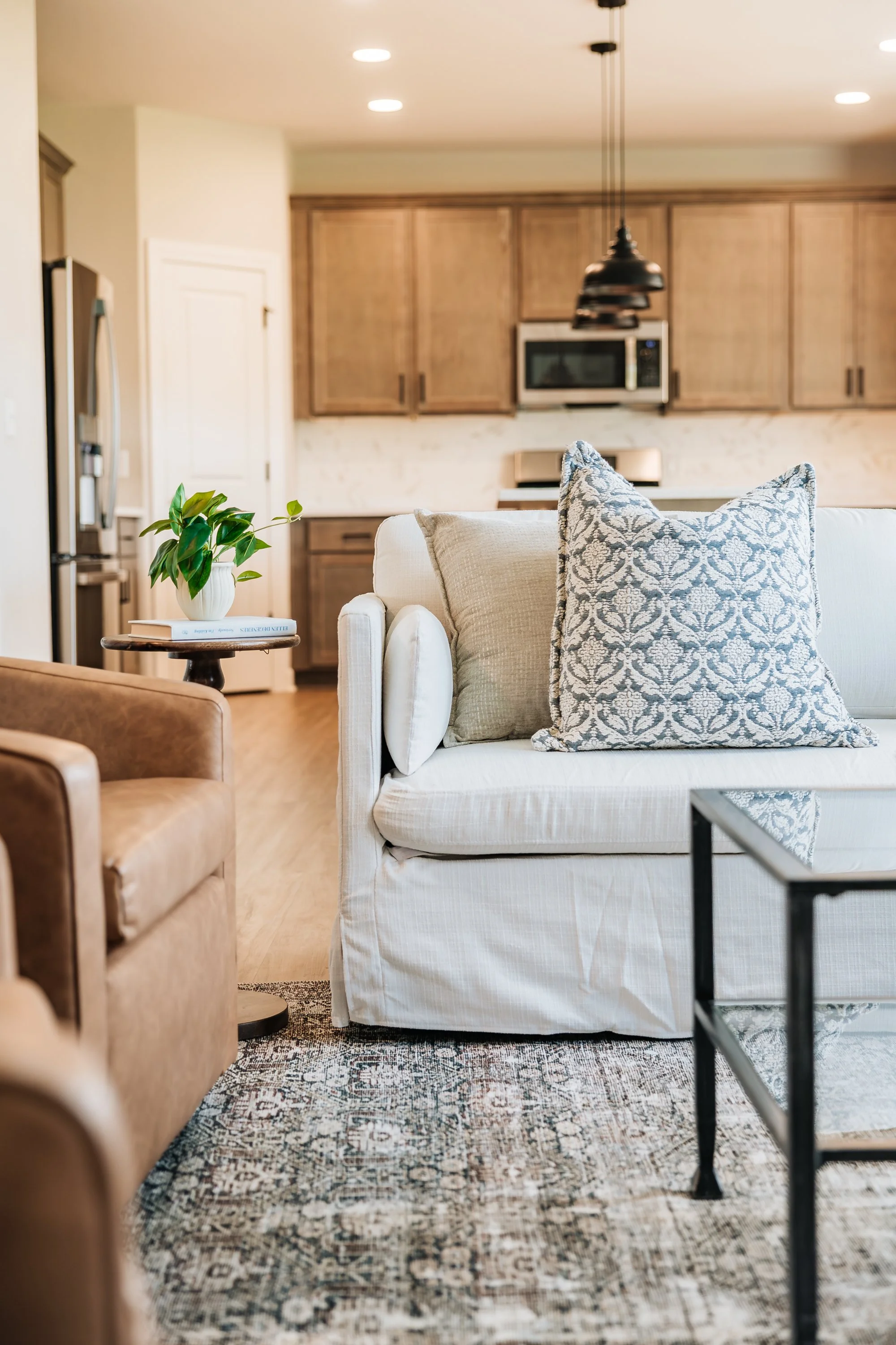 Living room with a white sofa, patterned pillows, a brown armchair, and a small side table with a green plant in a white pot, with a view of a kitchen in the background.