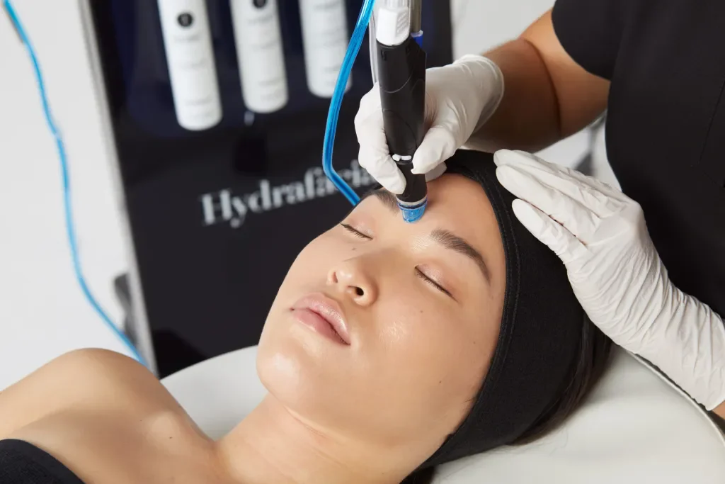 A woman receiving a facial treatment with a skincare device on her forehead while lying down with eyes closed, in a professional clinic setting.