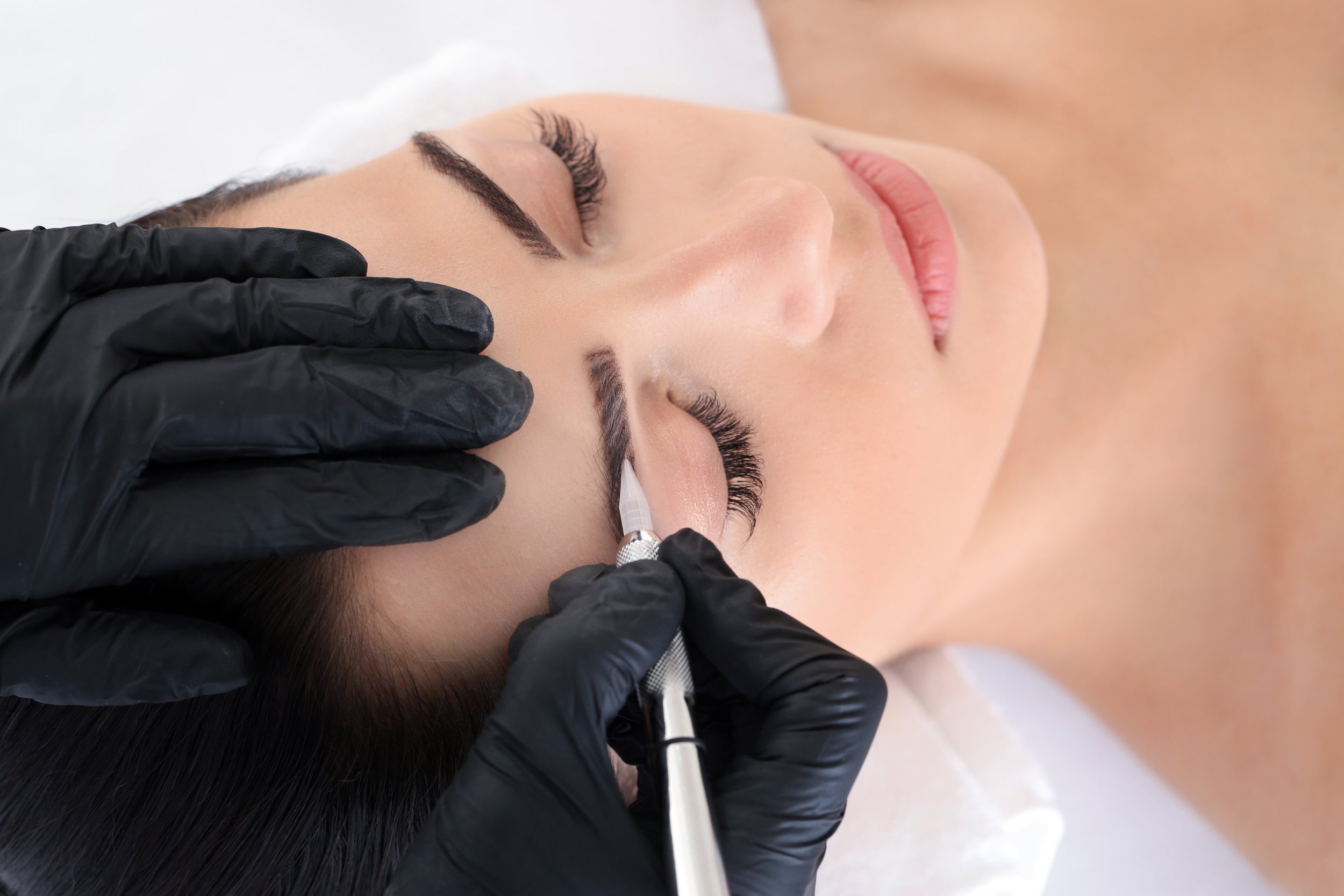 Woman receiving semi-permanent eyebrow tattooing or microblading procedure by a technician wearing black gloves, with a small tool near her eyebrow.